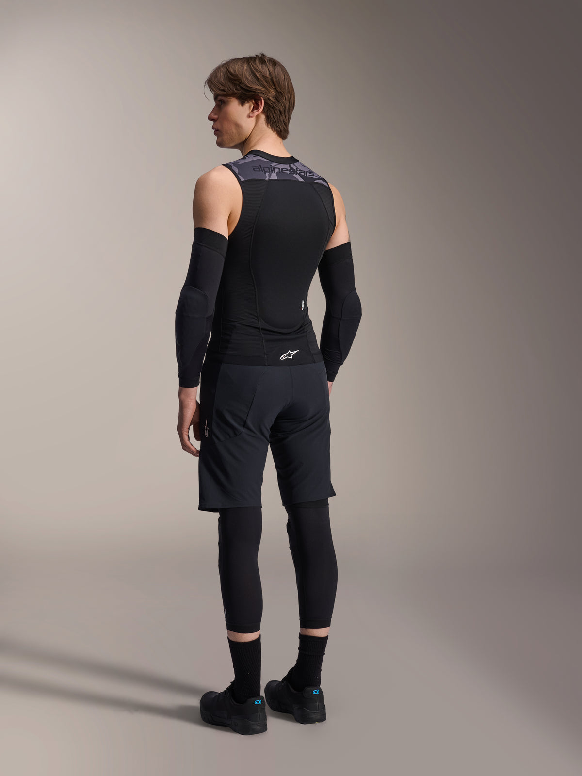 Modèle masculin de dos portant le gilet de protection Alpinestars A-Motion Plasma, Black Camo, design sans manches en maille avec protection dorsale intégrée, motif camouflage gris sur les épaules, porté avec des coudières noires et un short de VTT, équipement de cyclisme professionnel