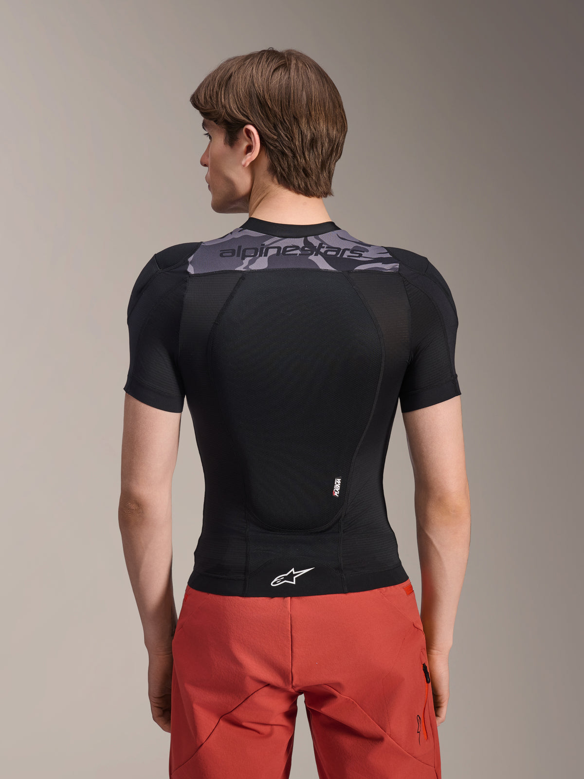 Veste de protection Alpinestars A-Motion Plasma - Manches courtes, noir avec accents camouflage gris sur le haut du dos, vue arrière montrant la protection dorsale intégrée et le logo Astars blanc à l'ourlet, portée avec un short rouge, équipement technique de protection pour VTT