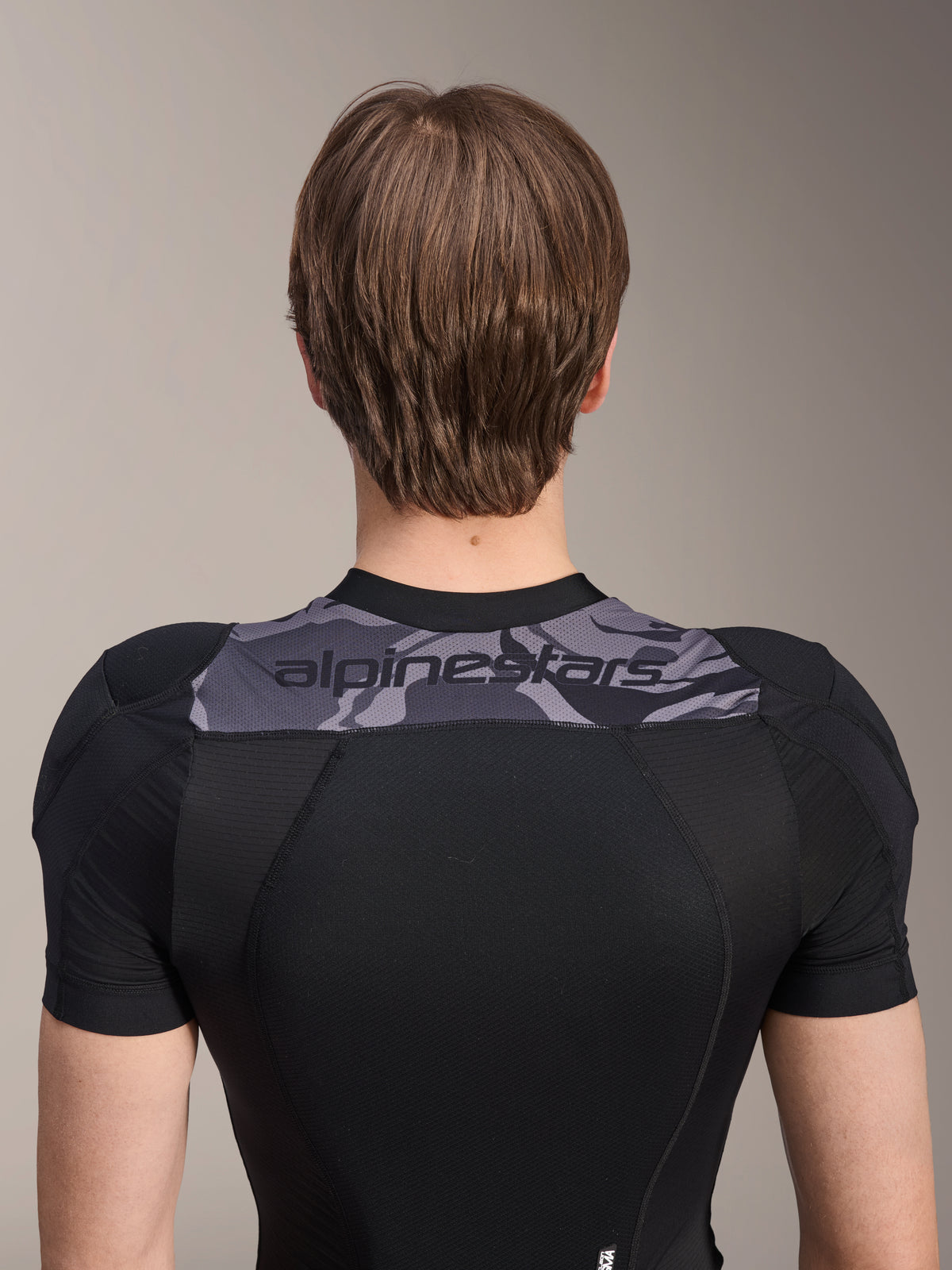 Veste de protection Alpinestars A-Motion Plasma - Manches courtes, vue arrière du haut du torse, noire avec empiècement camouflage gris sur les épaules arborant le logo Alpinestars, construction en maille extensible respirante pour la protection VTT