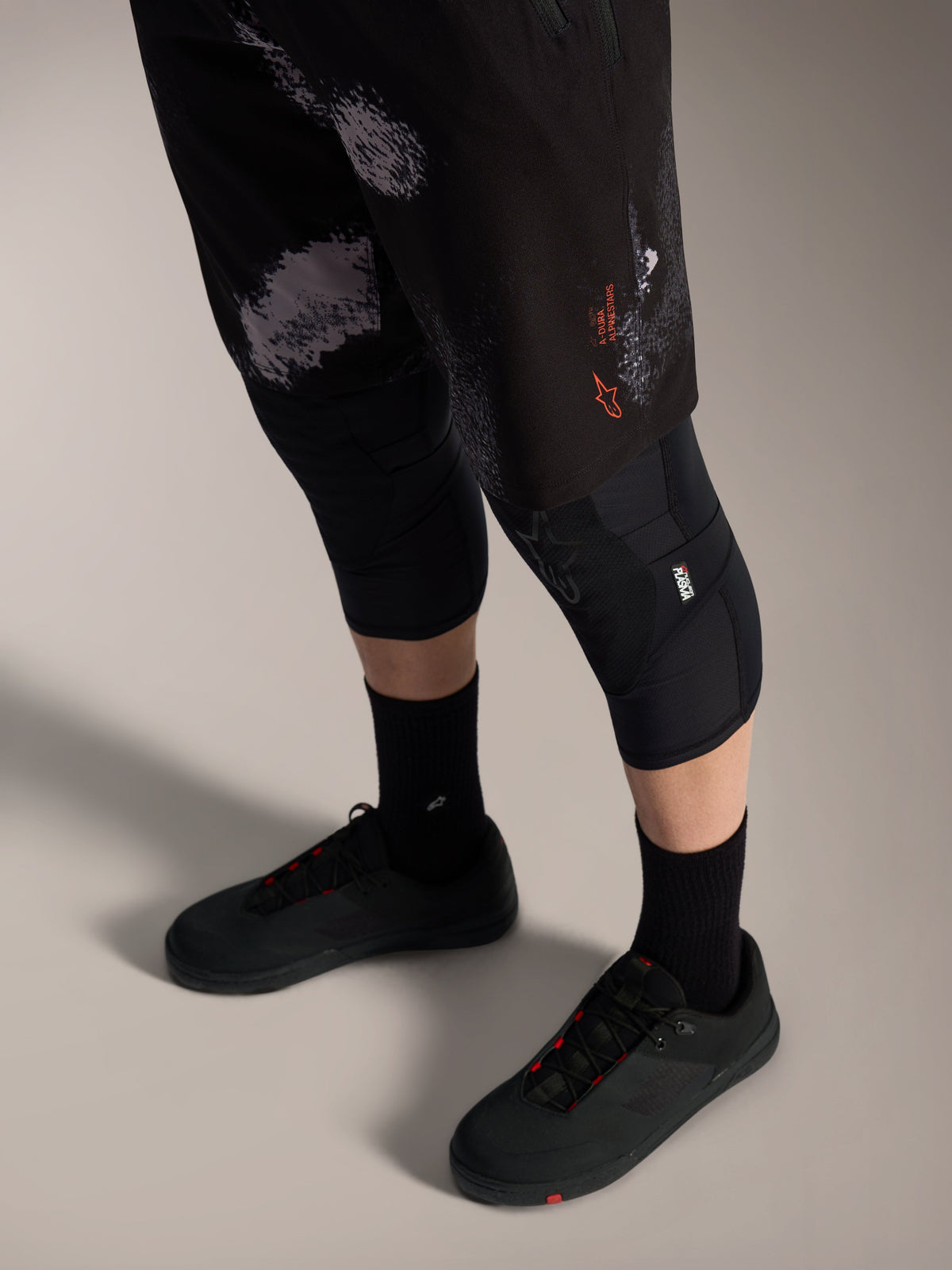Shorts Alpinestars A-Dura Ride Black Camo, équipement VTT avec logo orange, genouillères et chaussures de vélo.