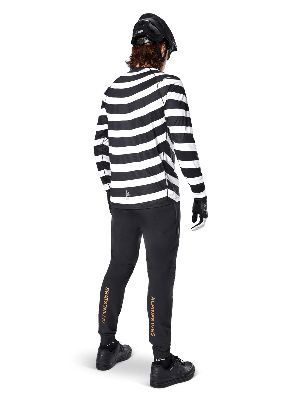 Pantalon Alpinestars A-Aria Elite, pantalon de VTT noir, avec logo vertical doré sur les mollets, porté par un cycliste avec un maillot à manches longues rayé noir et blanc, casque noir et chaussures noires, vue arrière montrant la coupe performance et un design prêt pour les sentiers.