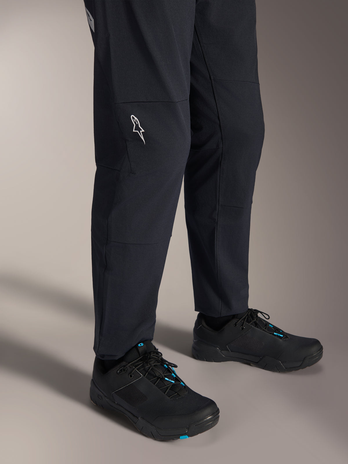 Pantalon Alpinestars A-Dura Elite, pantalon de VTT noir, vue du bas de la jambe avec logo Alpinestars blanc sur le genou, porté avec des chaussures de cyclisme noires et bleues, construction durable pour la pratique du VTT