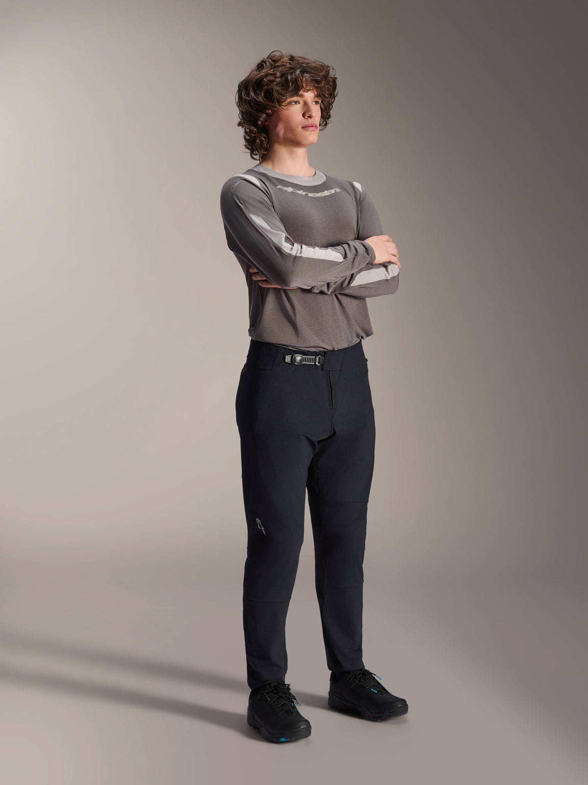 Modèle masculin portant le pantalon Alpinestars A-Dura Elite, pantalon de VTT noir, associé à un maillot gris à manches longues et des chaussures de cyclisme noires, système de fermeture à cliquet à l'avant, équipement professionnel pour le trail et l'enduro