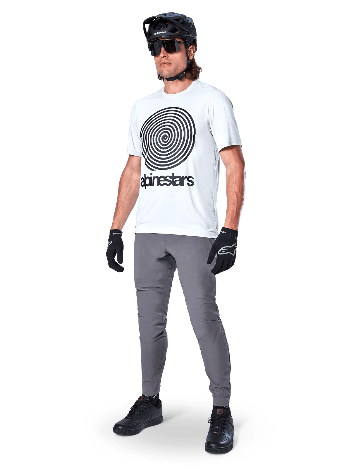 Pantalon Alpinestars A-Dura, pantalon de VTT, gris foncé, porté avec un t-shirt blanc à motif graphique en spirale, un casque de VTT noir, des lunettes de soleil et des gants noirs, textile extensible 2 voies durable pour le cyclisme tout-terrain