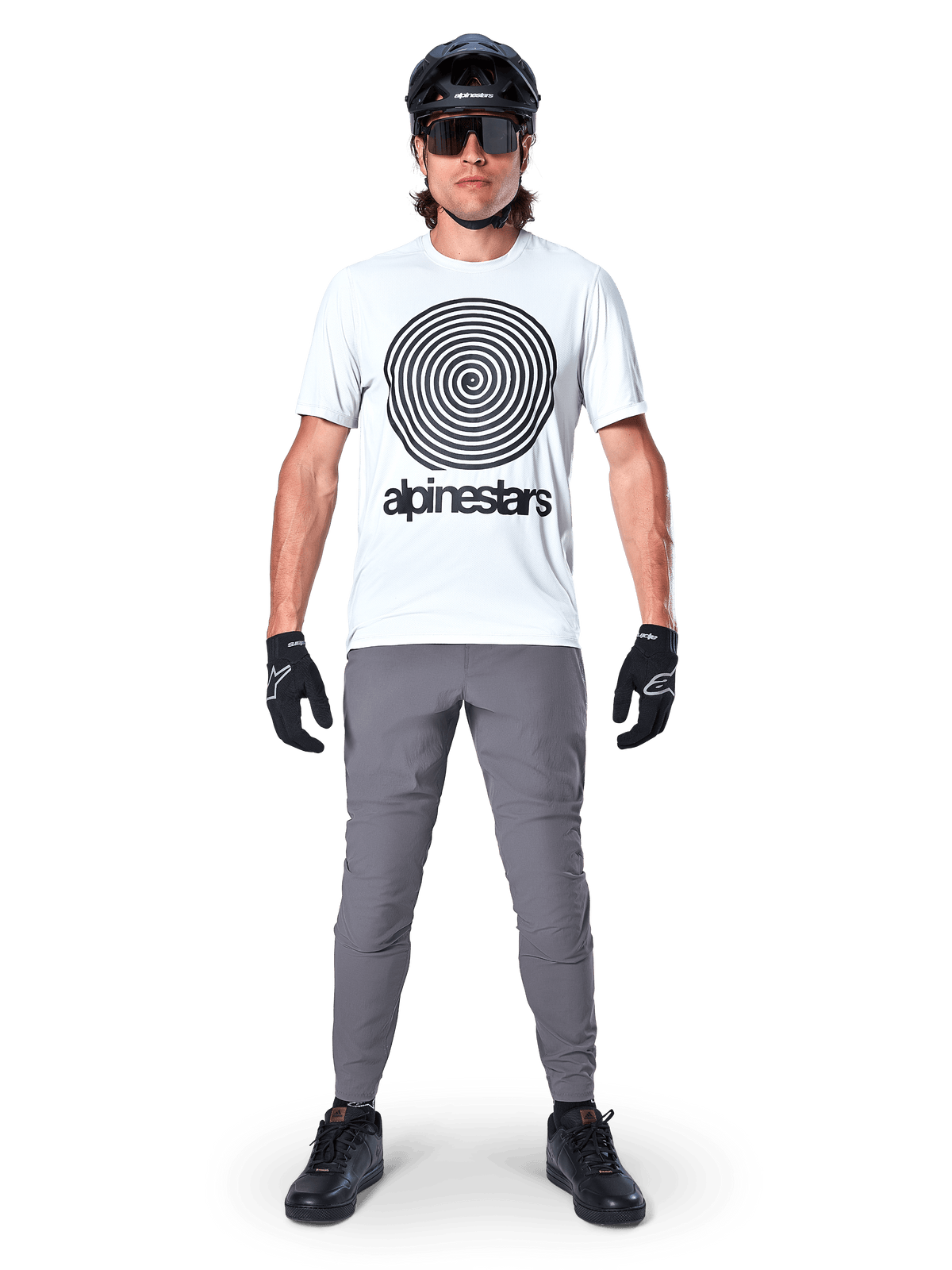 Pantalon Alpinestars A-Dura, pantalon de cyclisme VTT, gris foncé, porté avec un t-shirt blanc à motif graphique, un casque noir et des gants, doté d'une conception extensible 2 voies durable et d'une coupe régulière pour le VTT