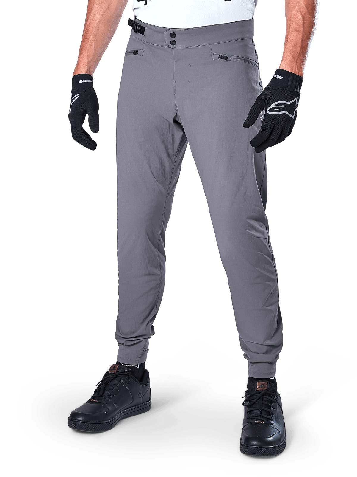 Alpinestars A-Dura Pants, pantalon de VTT gris foncé, doté d'une double fermeture à pression à la taille et de poches zippées, bas de jambes élastiqués, porté avec des gants de cyclisme noirs et des baskets noires