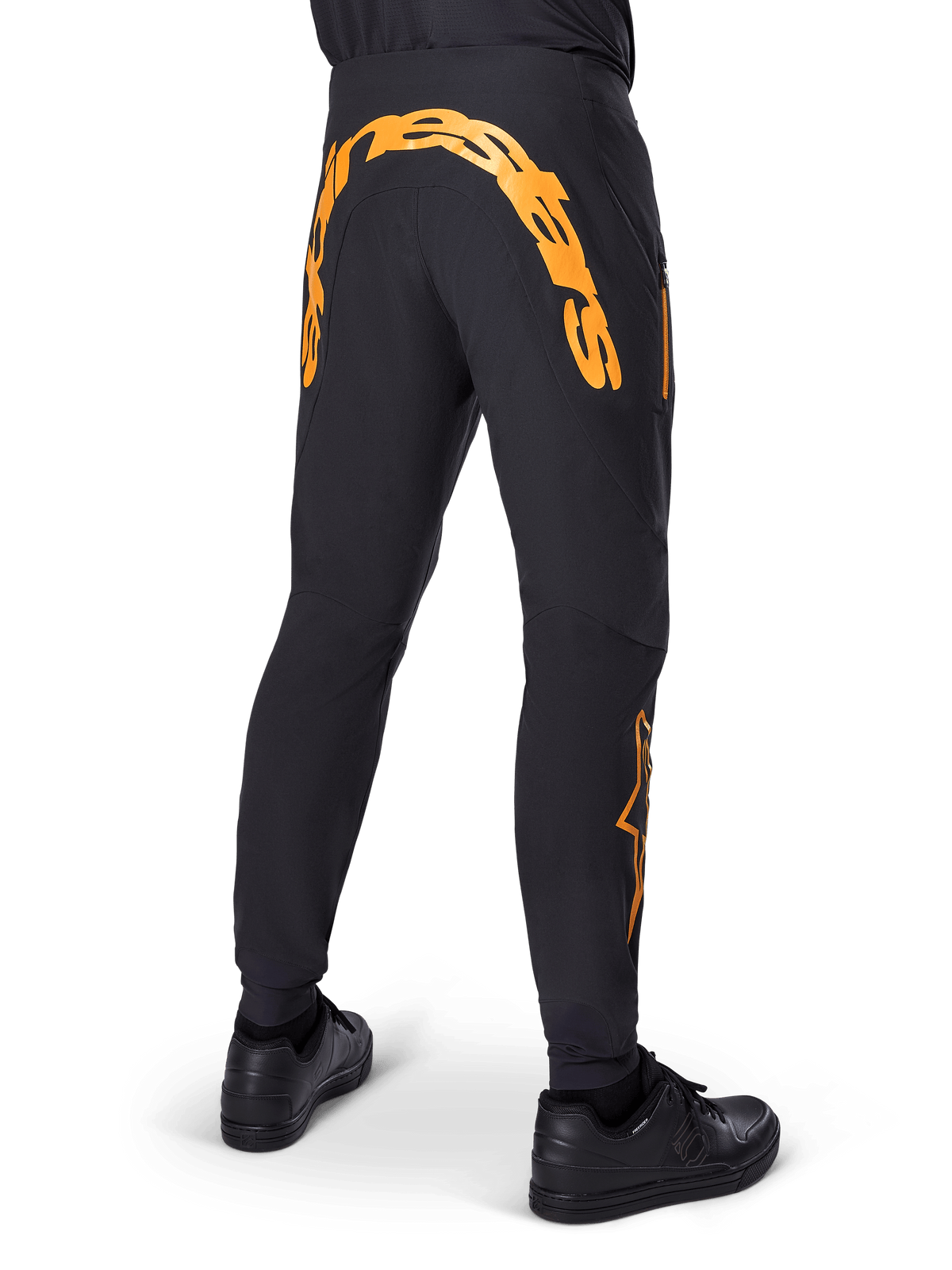 Pantalon Alpinestars A-Supra Race, pantalon de VTT noir, vue arrière avec un grand lettrage Alpinestars orange sur l'empiècement arrière, logo orange sur le bas de la jambe, coupe performance avec poche zippée, porté avec des chaussures noires