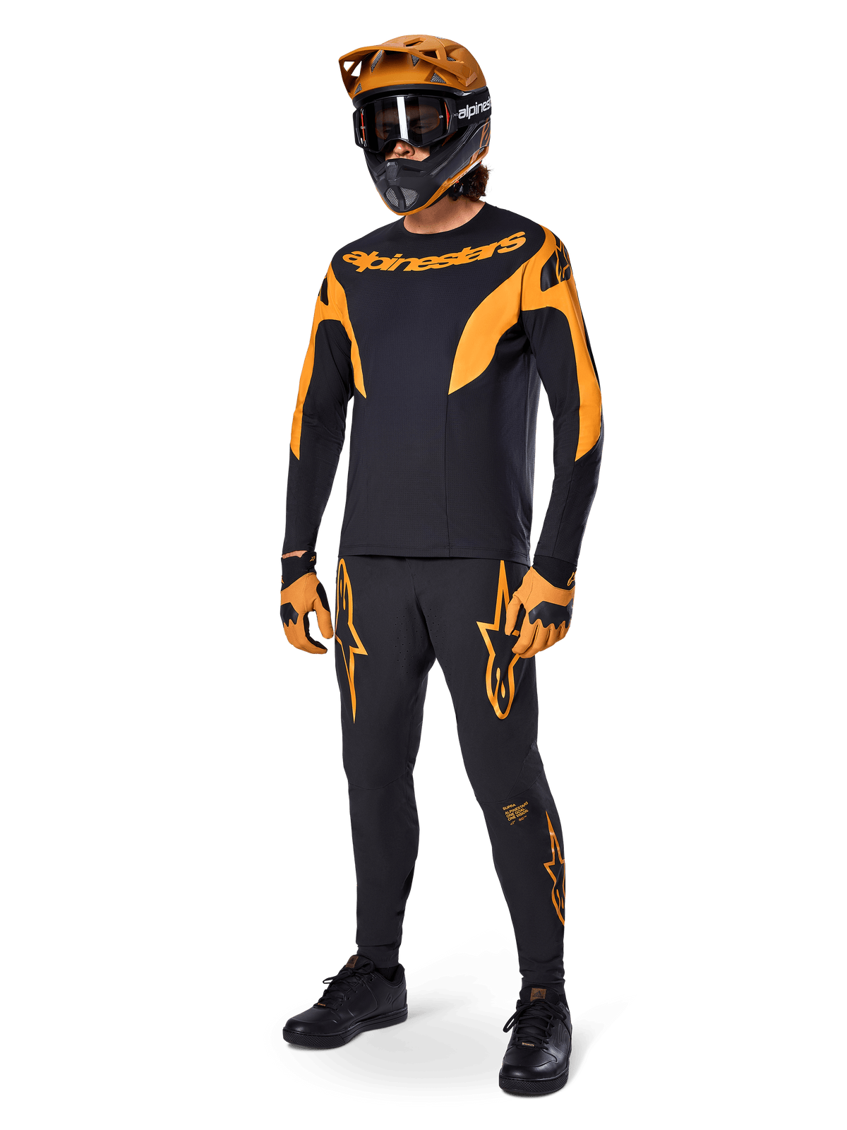 Pilote de VTT portant une tenue de course complète, pantalon de course Alpinestars A-Supra noir avec des accents orange, maillot à manches longues noir et orange assorti, casque orange avec lunettes noires, gants orange et noir, et chaussures noires, design de kit de course VTT professionnel