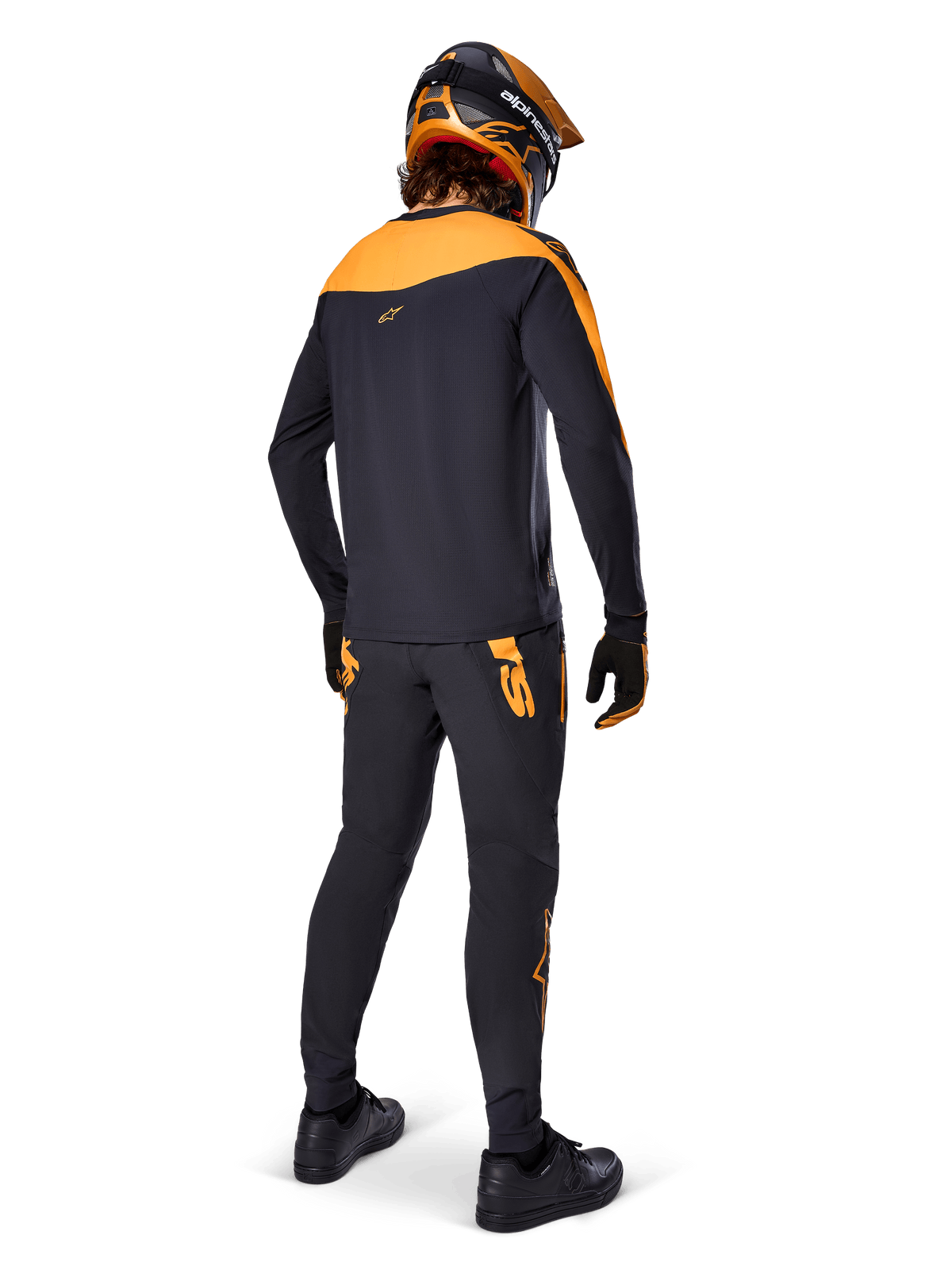Alpinestars A-Supra Race Pants, pantalon de course VTT, noir avec logos orange, vue de dos sur un pilote portant un maillot, un casque et des gants noir et orange assortis, mettant en avant le logo inspiré du MX sur l'empiècement arrière et la coupe performance pour les sentiers techniques.