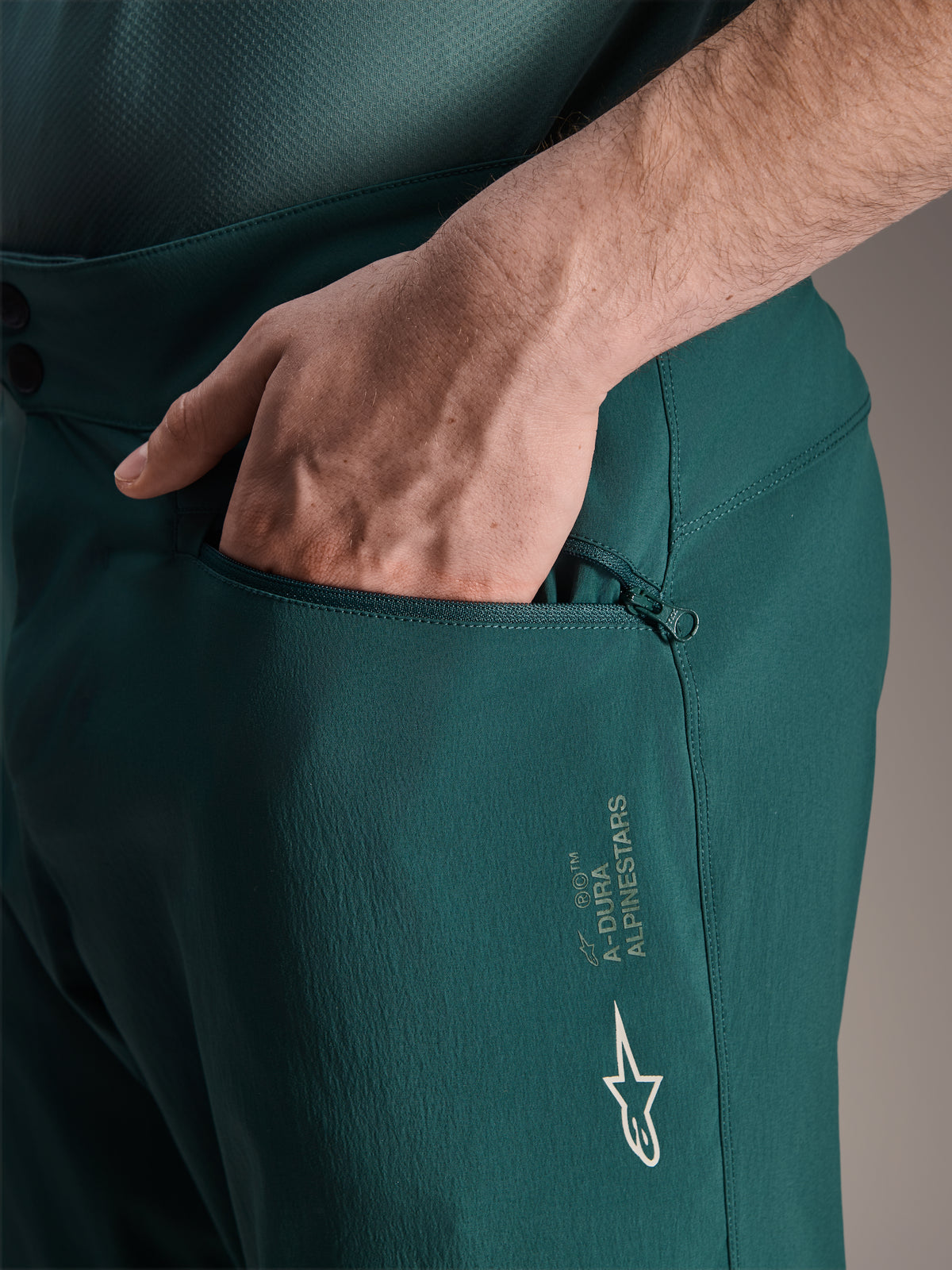 Short Alpinestars A-Dura en Dark Pine, vue rapprochée montrant une main dans la poche zippée doublée en maille, avec logo étoile Alpinestars blanc et marquage A-Dura, construction textile durable pour le VTT