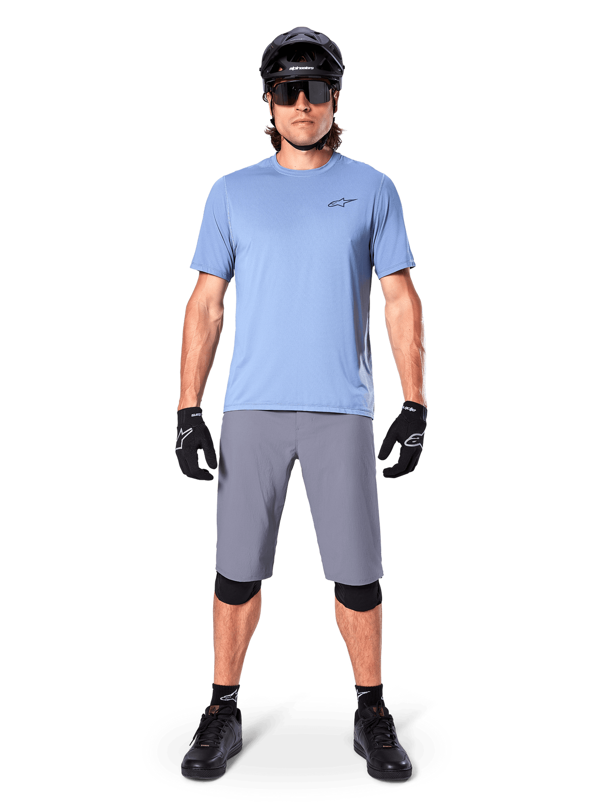 Male rider wearing Alpinestars A-Dura Shorts in Dark Gray, styled with a light blue short-sleeve t-shirt, black helmet, sunglasses, and black knee pads, complete mountain biking outfit for off-road performance.