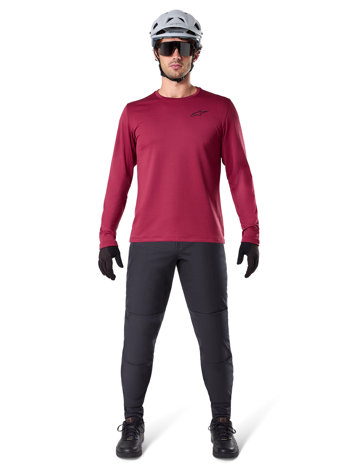 Modèle masculin portant le maillot thermique Alpinestars A-Dura bordeaux, haut de cyclisme isolé à manches longues, associé à un pantalon de VTT gris foncé, un casque blanc, des lunettes de soleil noires et des gants noirs, équipement professionnel de VTT pour temps froid