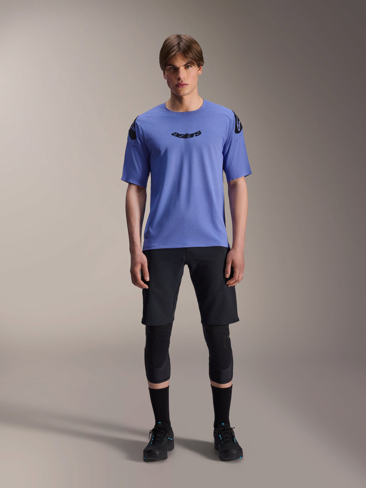 Modèle masculin portant le maillot à manches courtes Alpinestars A-Aria Polartec Elite en Baja Blue, logo Astars noir sur la poitrine, associé à un short de VTT noir, des genouillères et des chaussures de cyclisme noires, photo en studio pour équipement de VTT