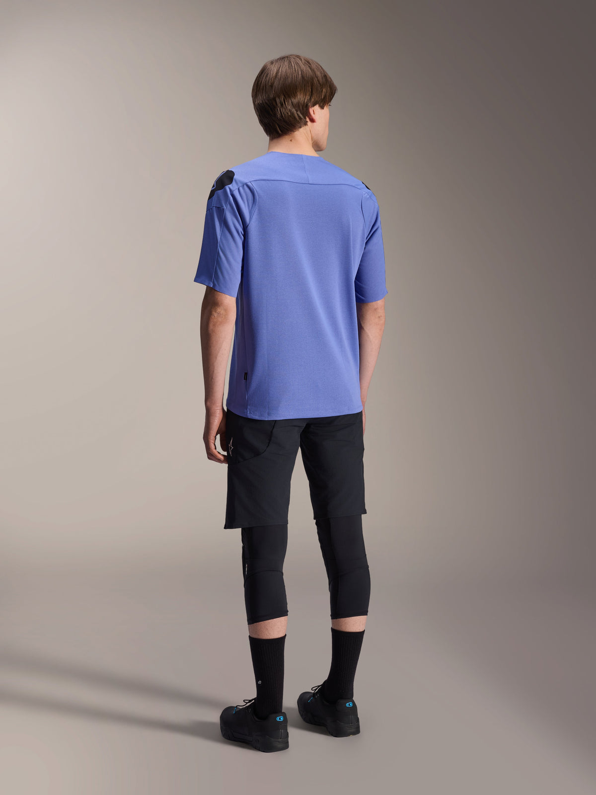Maillot à manches courtes Alpinestars A-Aria Polartec Elite en Baja Blue, vue de dos sur un modèle masculin, associé à un short de VTT noir et des protections de genoux, coupe performante et pan arrière allongé pour le trail.