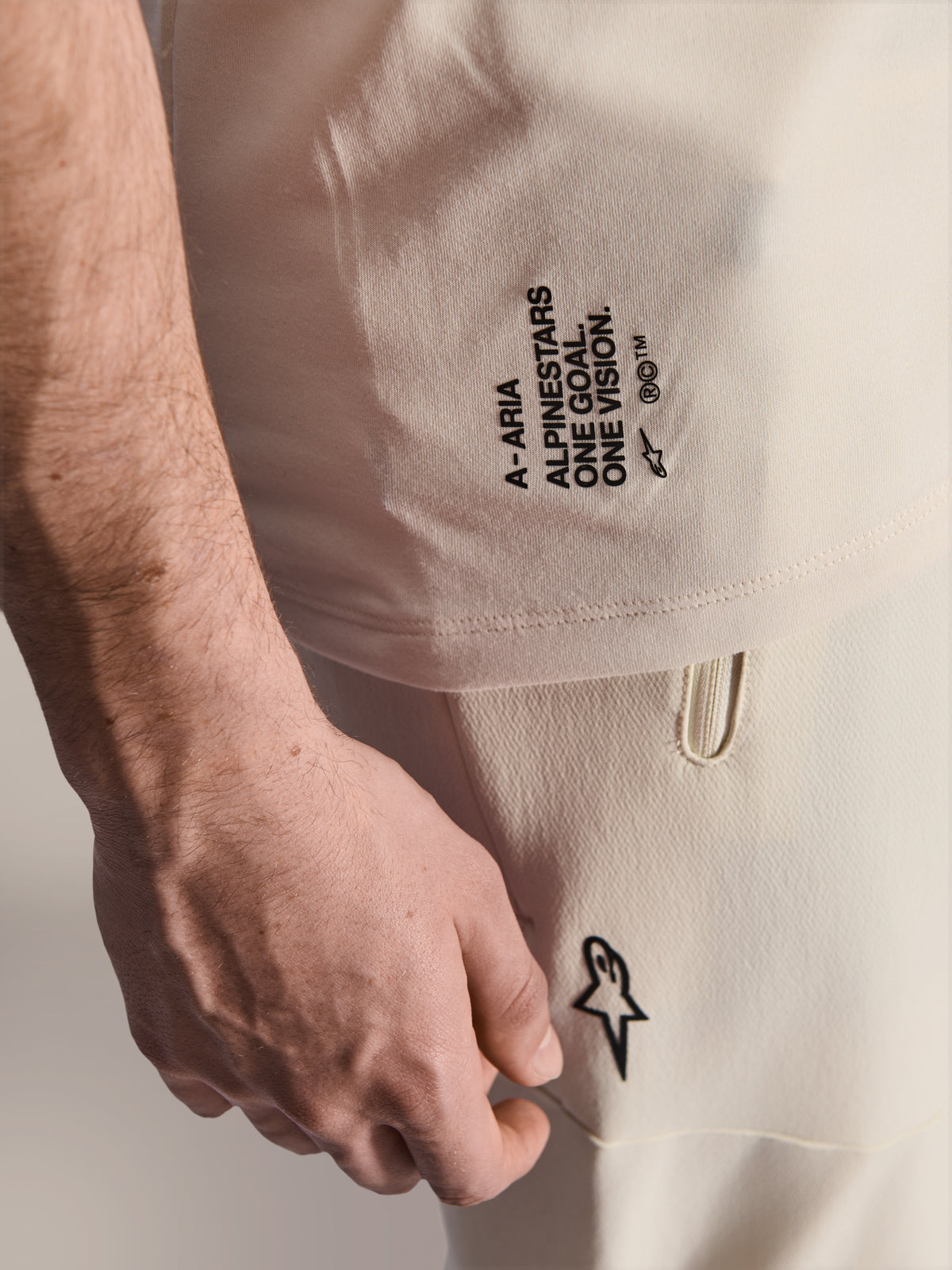 Alpinestars A-Aria Polartec Elite Short Sleeve Jersey, Ecru color, close-up of side branding featuring 'A-ARIA', 'ALPINESTARS', 'ONE GOAL. ONE VISION.', and Astars logo, worn with matching off-white pants, premium MTB performance apparel
