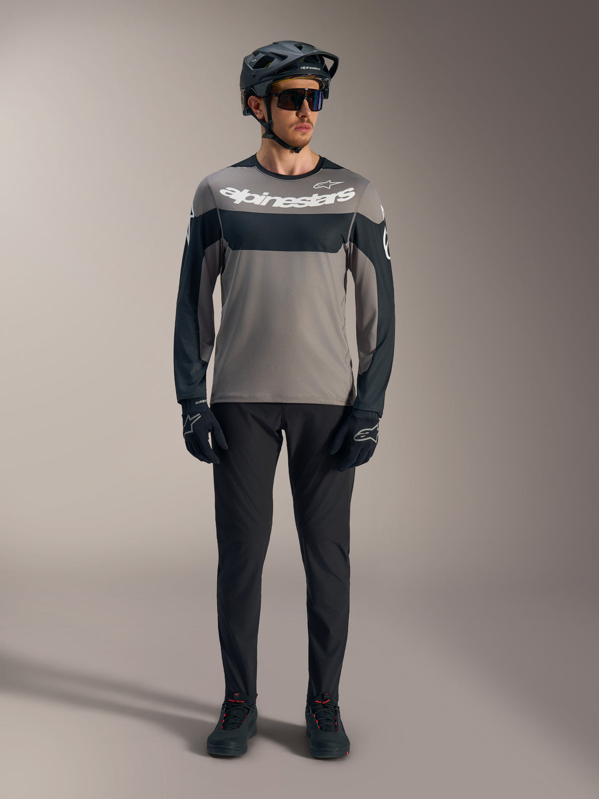 Maillot A-Dura Race à manches longues gris et noir, logo Alpinestars blanc, porté avec équipement VTT de performance.