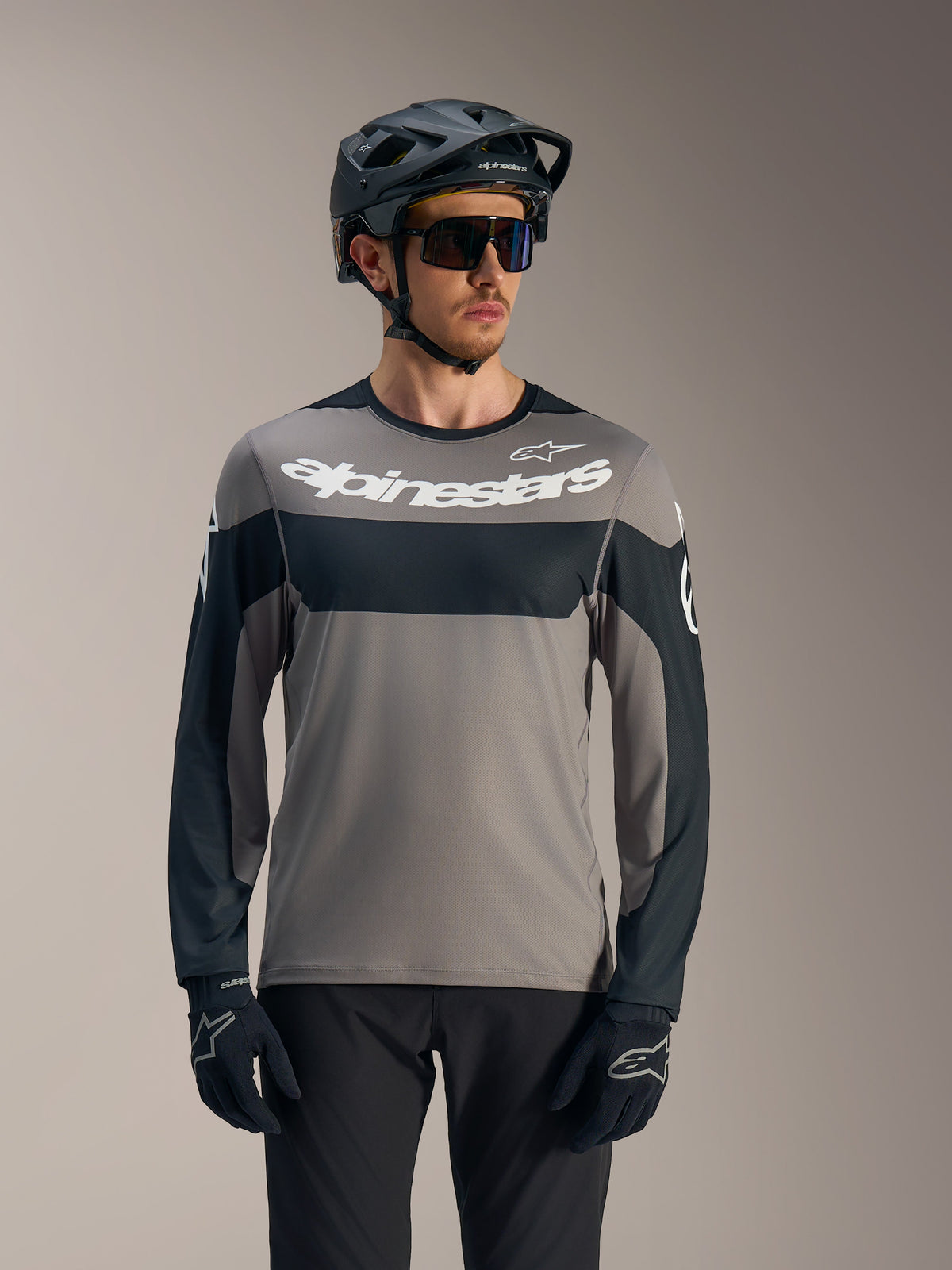 Maillot A-Dura Race manches longues gris et noir, logo blanc Alpinestars, porté avec un casque de VTT pour le trail.