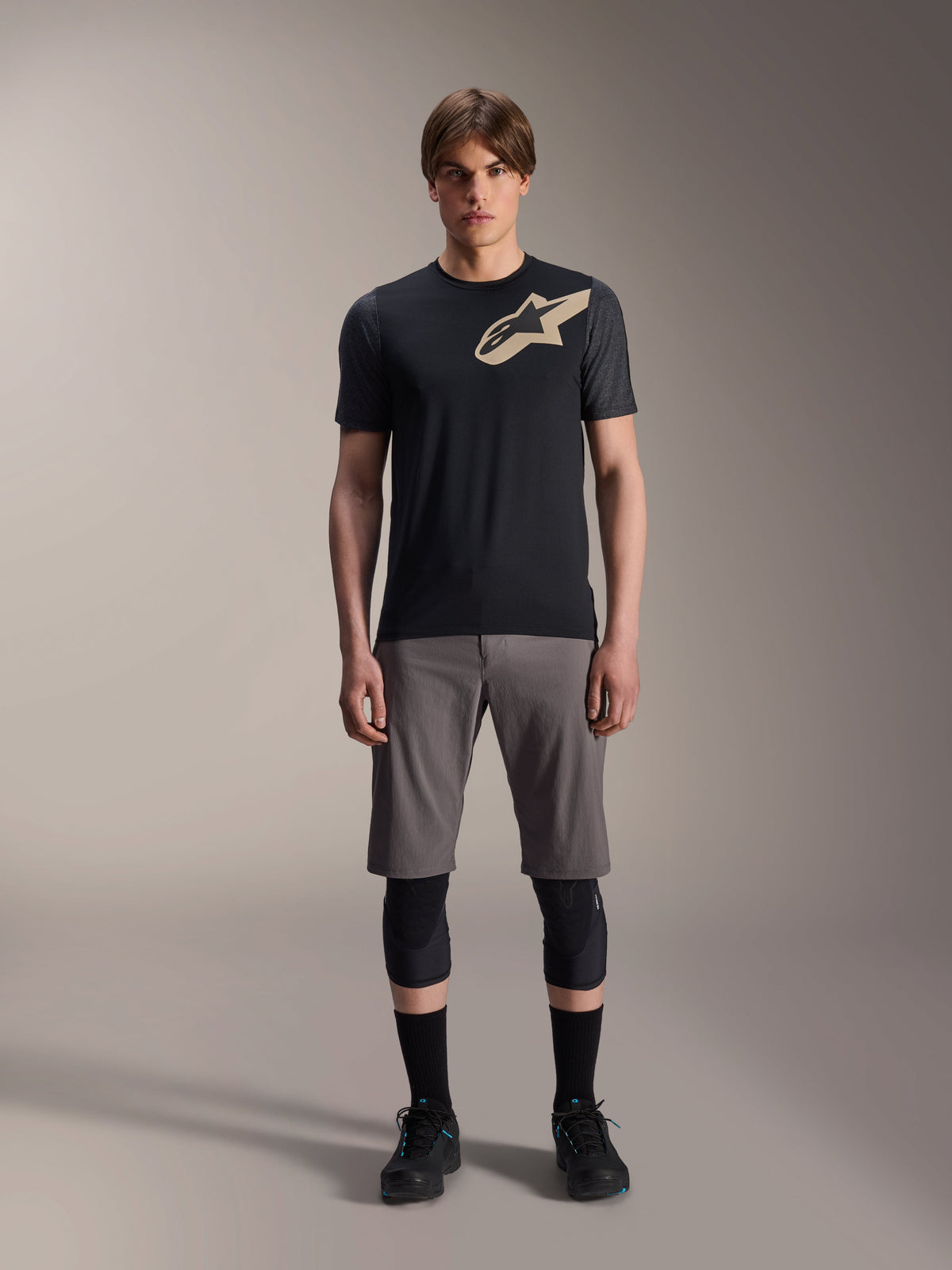 Alpinestars A-Dura Lipan Short Sleeve Jersey, Black with grey marl sleeves and tan Astars chest logo, worn by a male model with grey mountain bike shorts and black knee guards, performance MTB riding gear.