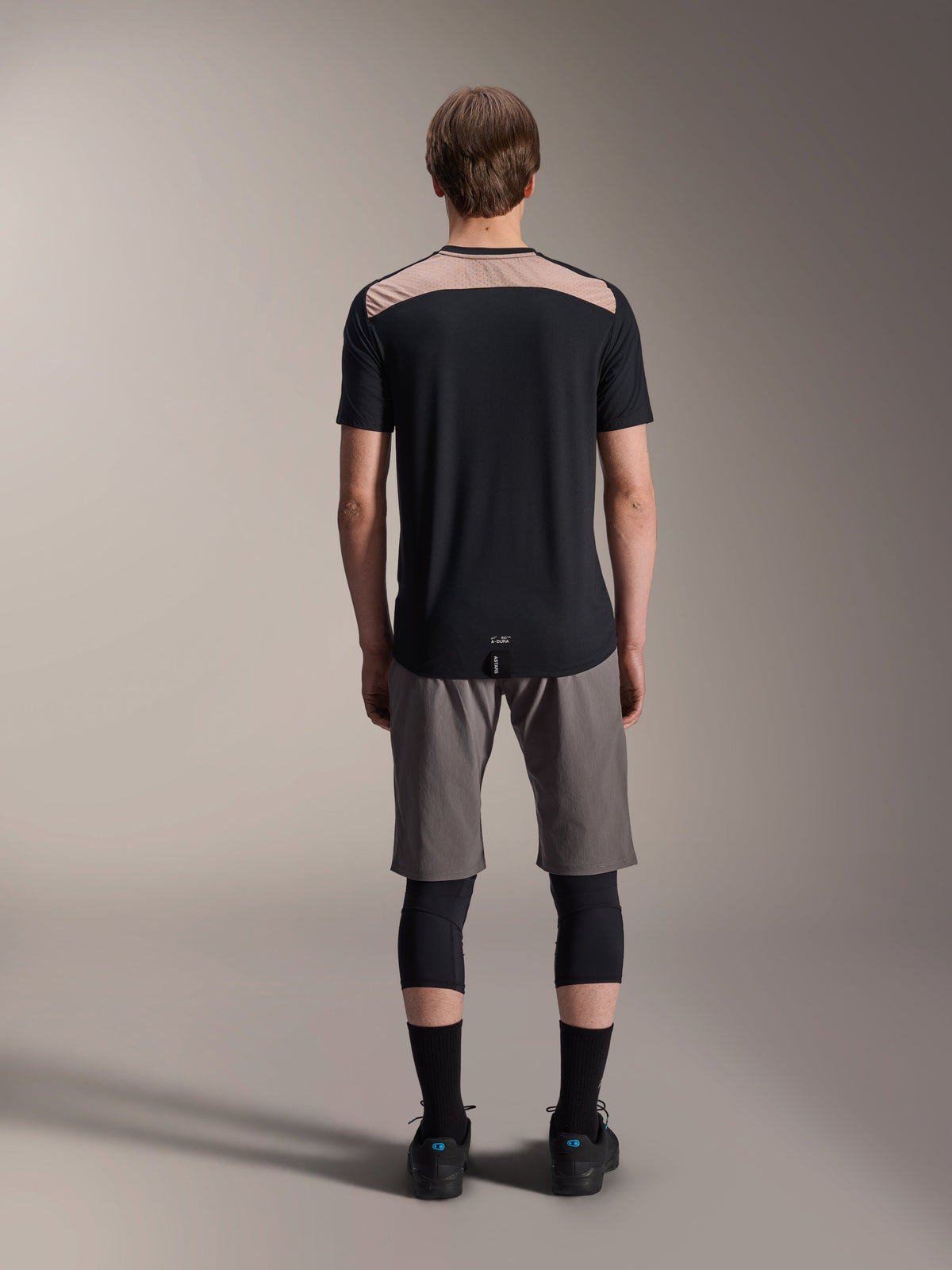Alpinestars A-Dura Lipan Jersey - Short Sleeve, Black with tan upper back panel, rear view showing dropped tail, worn with grey MTB shorts and black knee guards, performance mountain bike apparel