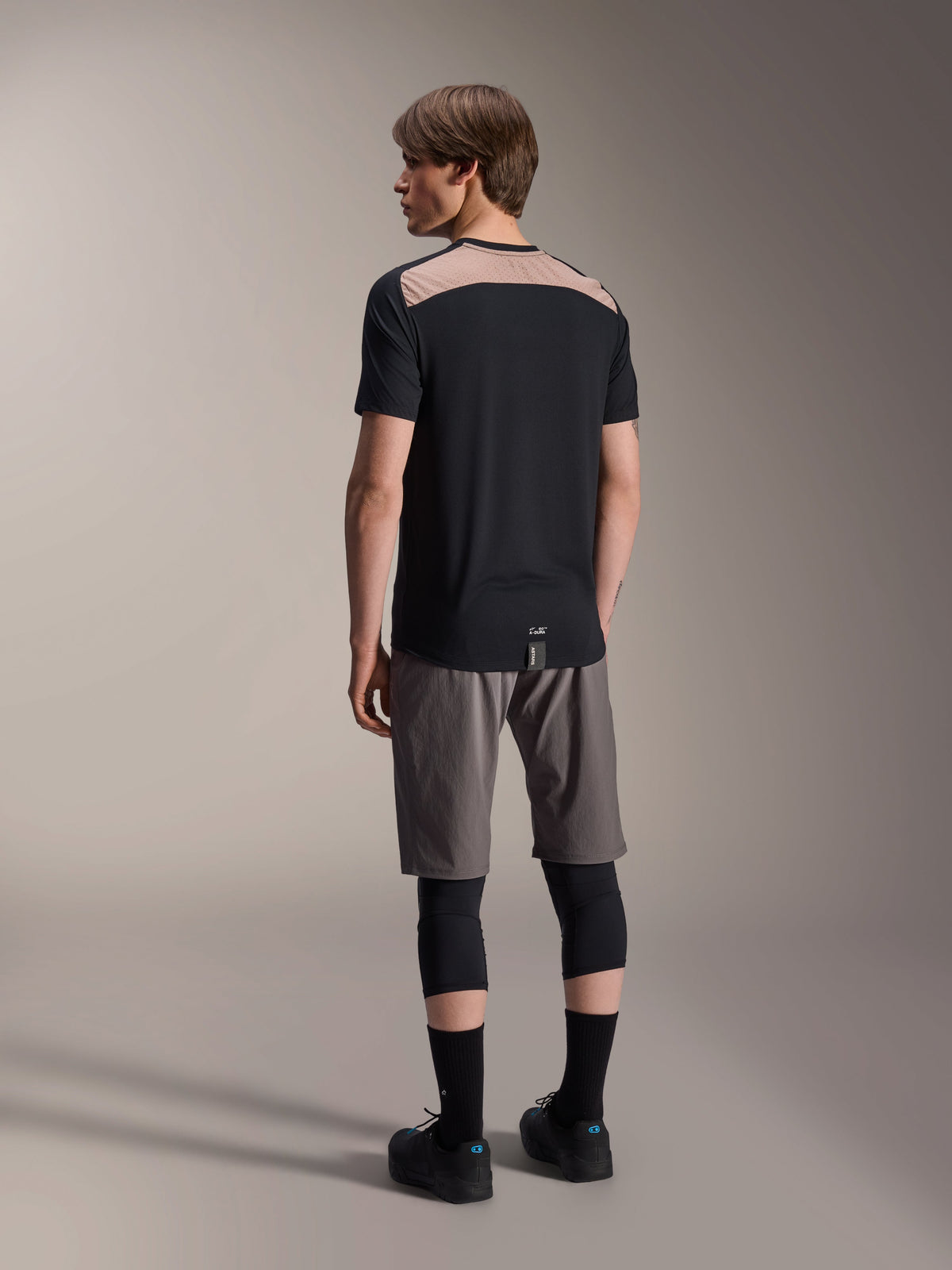 Alpinestars A-Dura Lipan Short Sleeve Jersey in Black, back view on model showcasing the light brown perforated mesh panel across the upper back, paired with grey mountain bike shorts and black knee guards for a complete trail riding outfit.