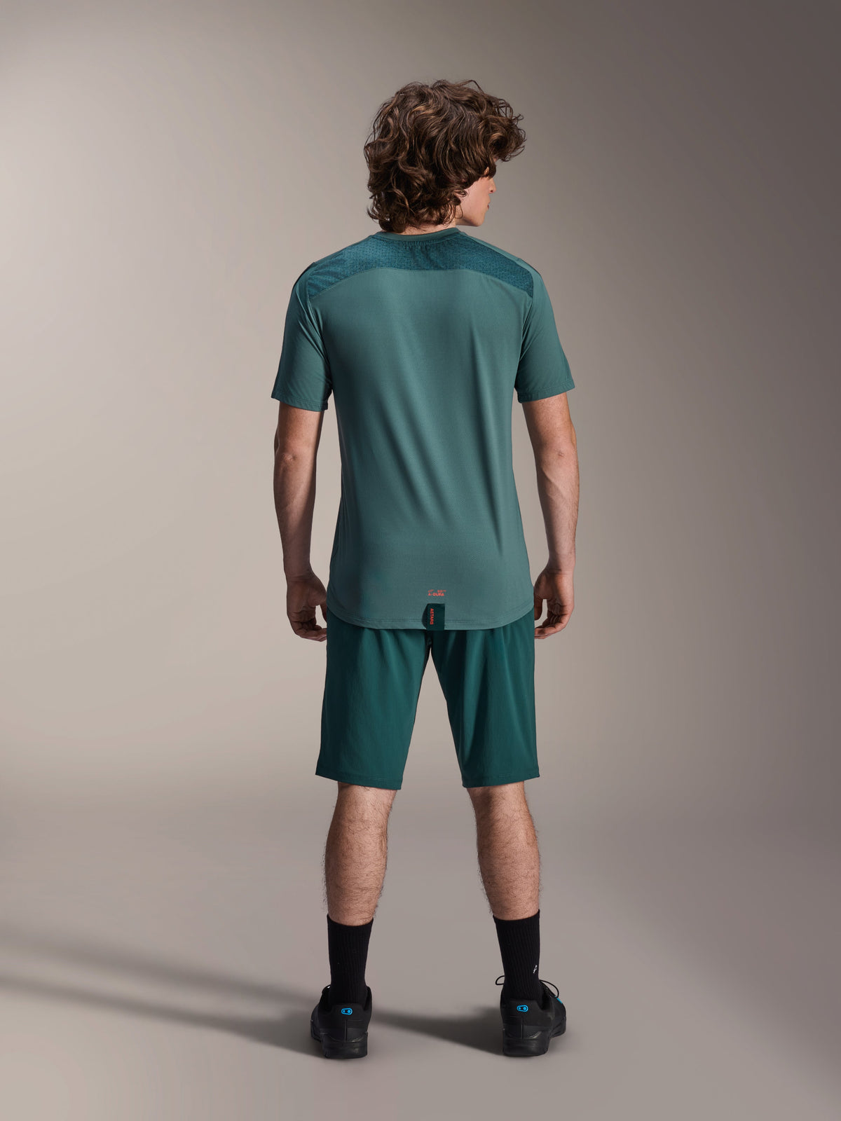 Alpinestars A-Dura Lipan Short Sleeve Jersey in Silver Pine, back view showing the textured jacquard shoulder panels and drop tail hem, worn with matching dark green mountain bike shorts and black footwear.
