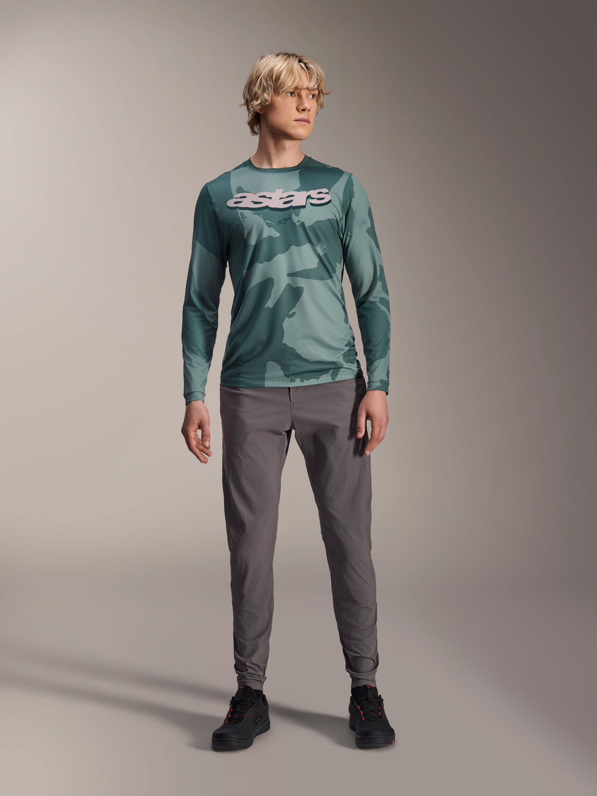 Maillot à manches longues Alpinestars A-Dura Hollow Camo, coloris Silver Pine Camo avec motif camouflage vert, grand logo « astars » blanc sur la poitrine, porté avec un pantalon technique gris et des chaussures de vélo noires, équipement VTT haute performance