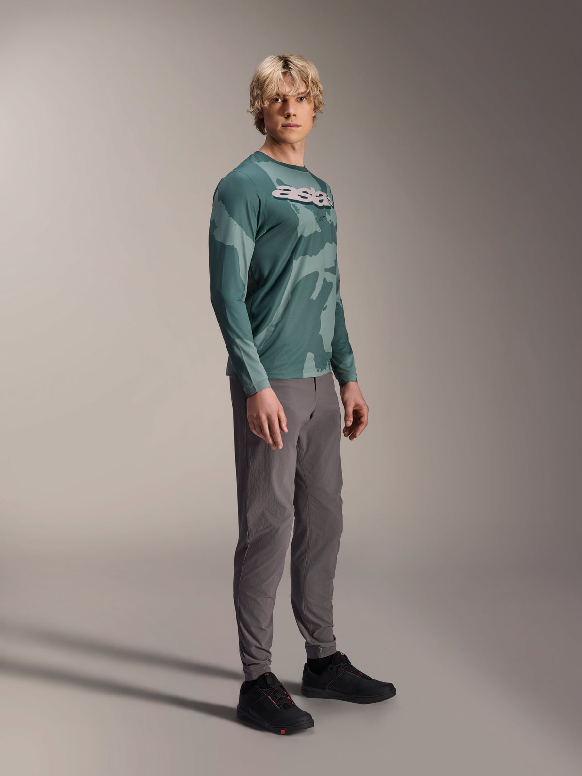 Maillot à manches longues Alpinestars A-Dura Hollow Camo, Silver Pine Camo, porté par un mannequin avec un pantalon gris et des chaussures noires, arborant un motif camouflage vert et un logo ASTARS blanc audacieux sur la poitrine, vêtement de VTT haute performance