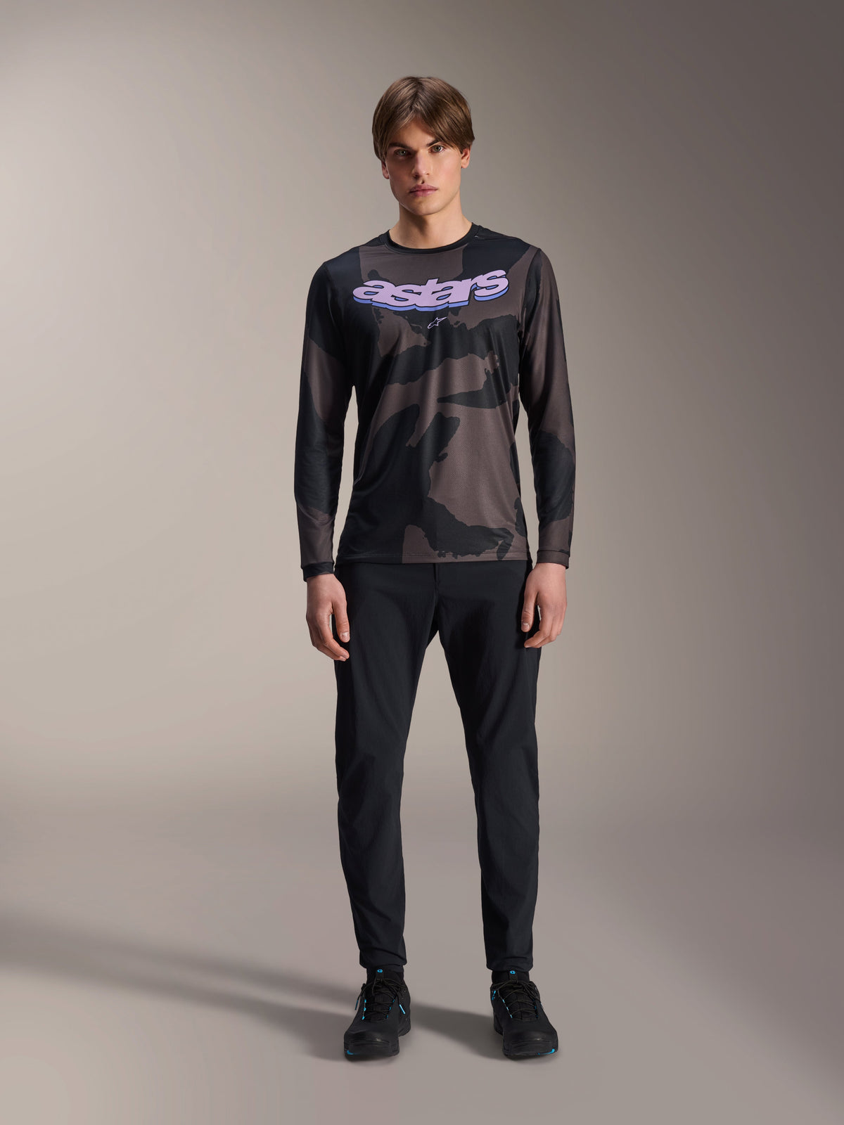 Modèle portant le maillot à manches longues Alpinestars A-Dura Hollow Camo en Black Camo, avec un logo 'astars' violet sur la poitrine, associé à un pantalon noir et des chaussures de cyclisme noires, équipement de VTT