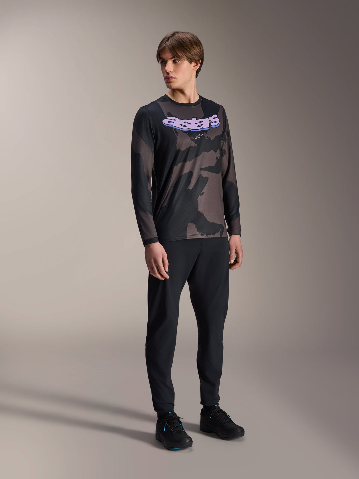 Modèle masculin portant le maillot à manches longues Alpinestars A-Dura Hollow Camo, motif Black Camo avec graphisme 'astars' violet sur la poitrine, associé à un pantalon technique noir et des chaussures noires, équipement de VTT décontracté
