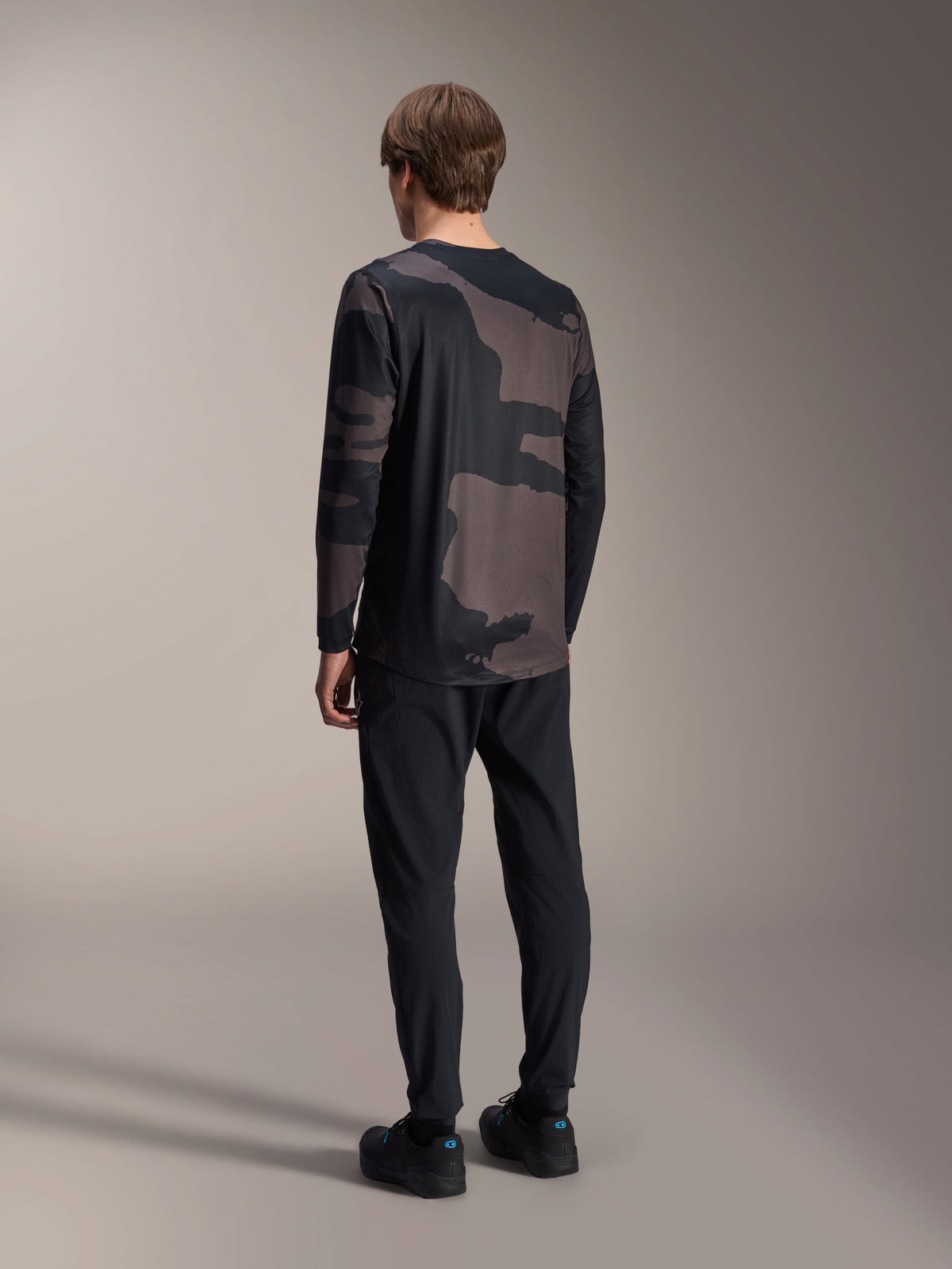 Maillot à manches longues Alpinestars A-Dura Hollow Camo, Black Camo, vue arrière du mannequin, maillot technique de VTT avec pan arrière allongé, associé à un pantalon noir et des chaussures de cyclisme, équipement de performance tout-terrain décontracté