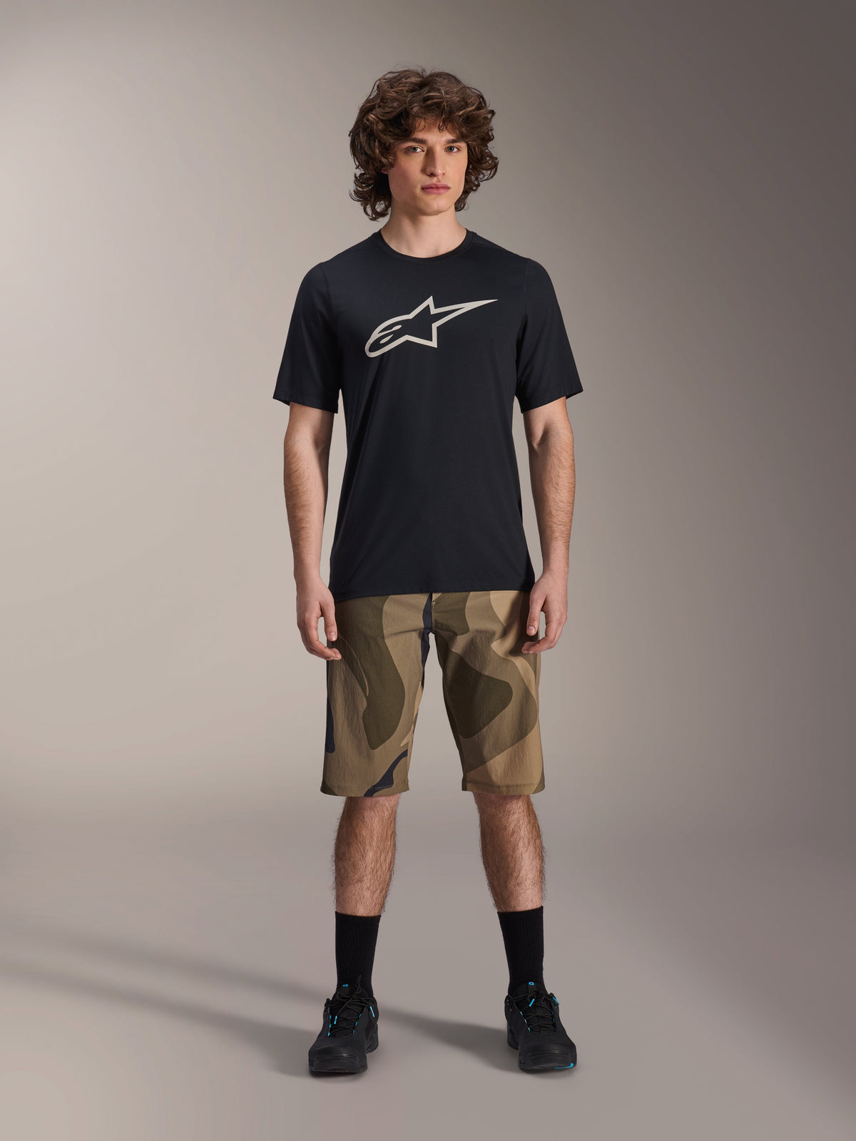 Alpinestars A-Dura Astar Short Sleeve Jersey, Black with large white Astars logo on the chest, worn by a model with camouflage MTB shorts and black cycling shoes, casual mountain bike apparel.