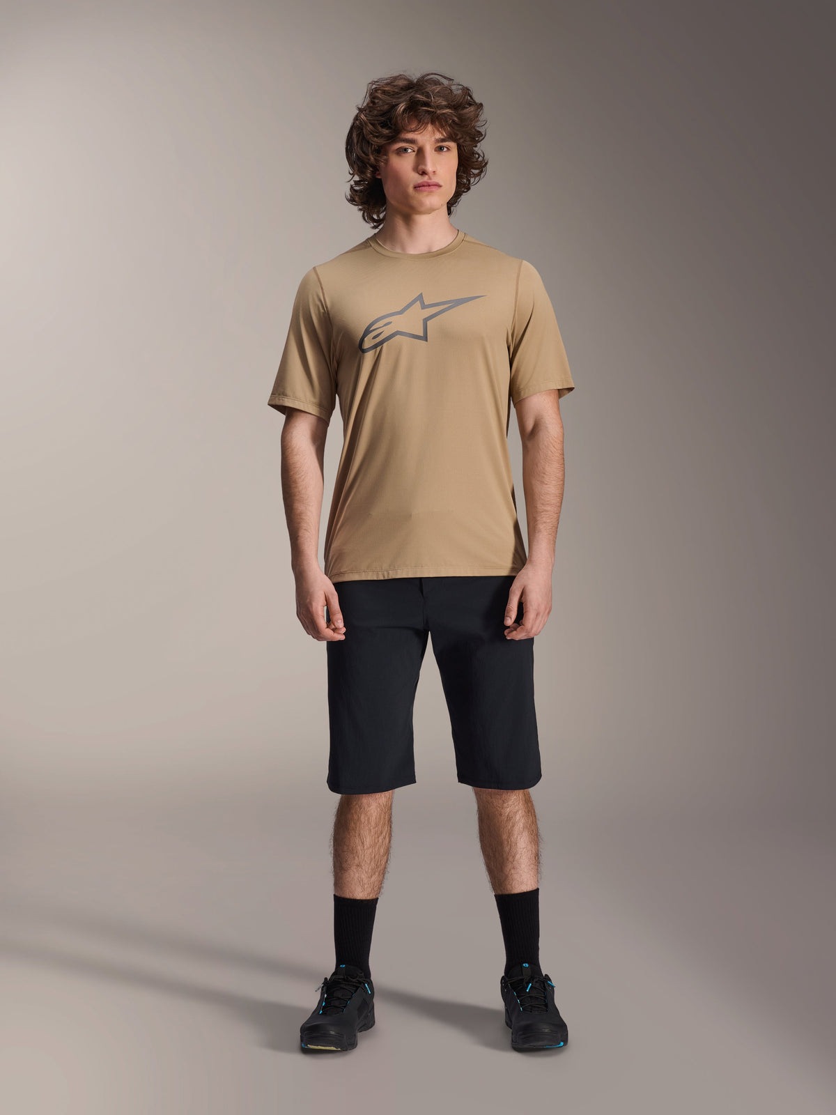 Maillot Alpinestars A-Dura Astar à manches courtes, marron clair, porté avec un short noir et des chaussures noires, arborant un grand logo Astar détouré sur la poitrine, tissu jacquard technique pour la pratique du VTT trail