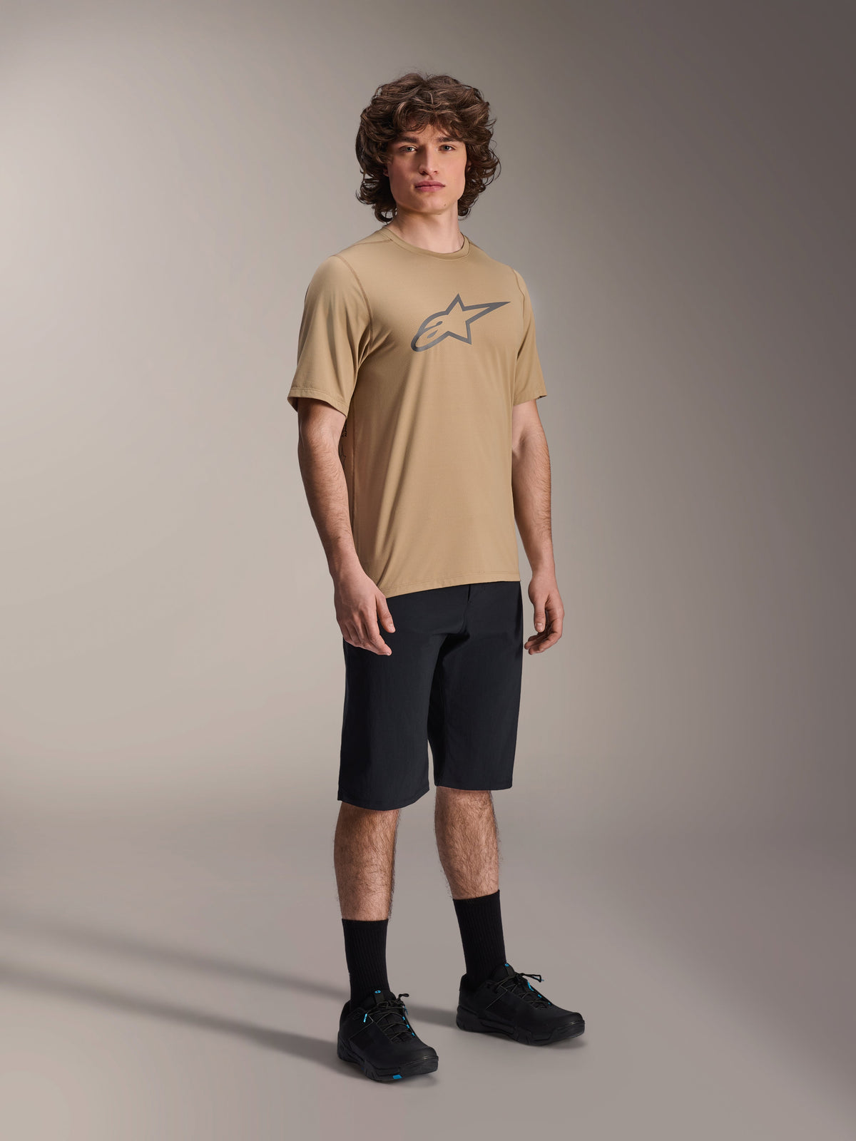 Maillot à manches courtes Alpinestars A-Dura Astar, marron clair, avec un grand logo Astar gris foncé sur la poitrine, porté par un mannequin avec un short de VTT noir, des chaussettes noires et des chaussures noires, tenue de VTT performante et décontractée dans un studio