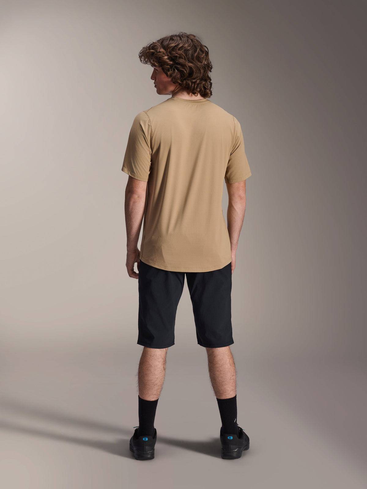 Cycliste masculin vu de dos portant le maillot à manches courtes Alpinestars A-Dura Astar en marron clair, associé à un short de VTT noir et des chaussettes noires, style de randonnée VTT décontracté.
