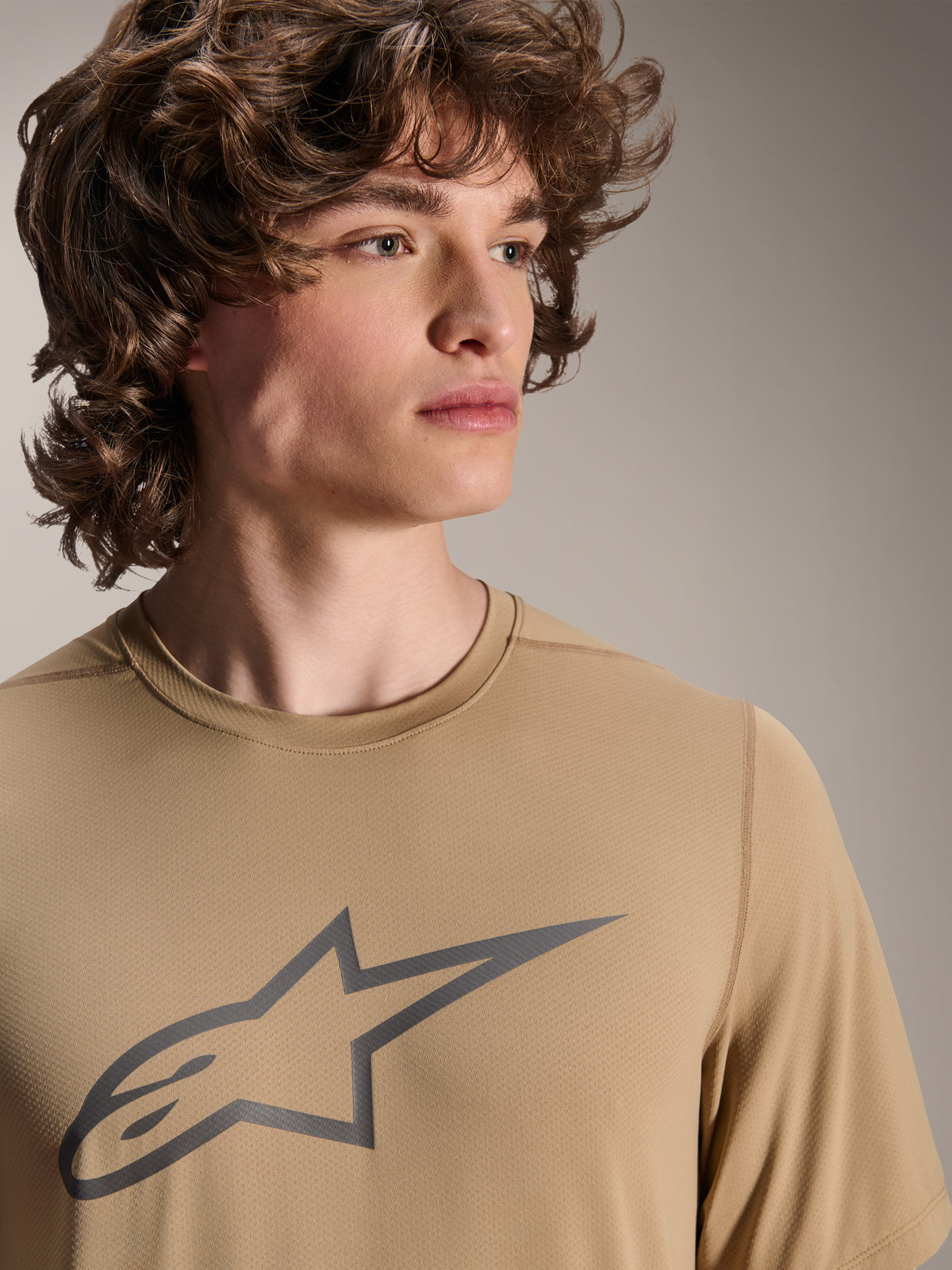 Modèle portant le maillot à manches courtes Alpinestars A-Dura Astar, maillot de VTT marron clair, avec un logo étoile gris sur la poitrine, tissu jacquard évacuant l'humidité, style cycliste décontracté