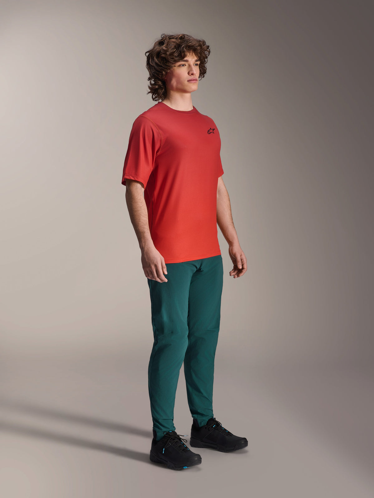 Alpinestars A-Dura Rise Jersey, Short Sleeve, Spicy Orange, featuring a small black chest logo, worn with dark teal pants and black shoes, lightweight moisture-wicking mountain bike apparel
