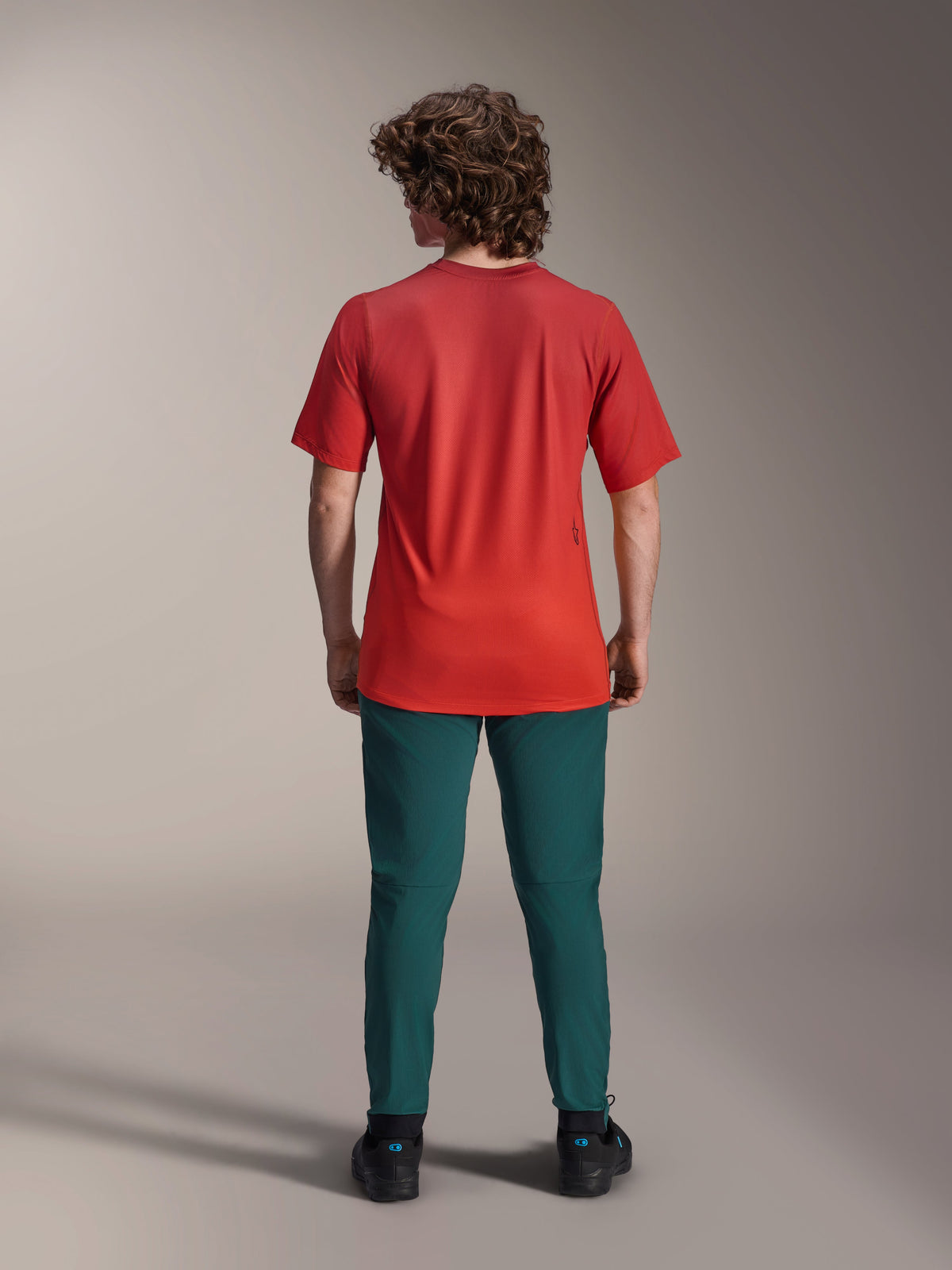 Alpinestars A-Dura Rise Jersey - Short Sleeve, Spicy Orange, rear view on a model wearing dark teal pants and black shoes, featuring a dropped tail and a small screen-printed logo on the back, lightweight performance MTB gear.
