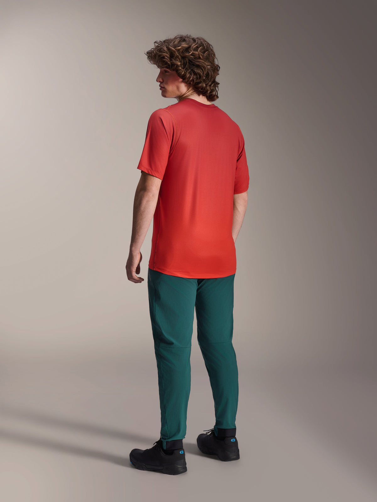 Alpinestars A-Dura Rise Jersey - Short Sleeve in Spicy Orange, back view of model wearing the cycling jersey with dark green mountain bike pants and black shoes, featuring a longer drop tail and performance fit for off-road riding