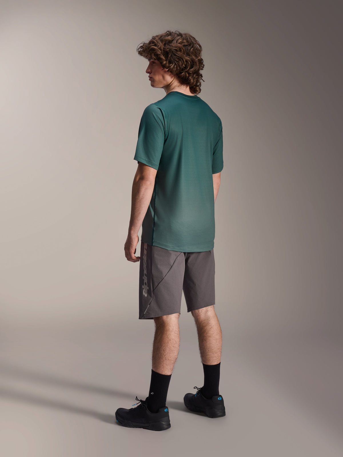 Alpinestars A-Dura Rise Jersey, Short Sleeve MTB Jersey, Silver Pine Green, worn with gray shorts and black shoes, rear view showing longer drop tail and regular fit, lightweight moisture-wicking fabric for trail riding