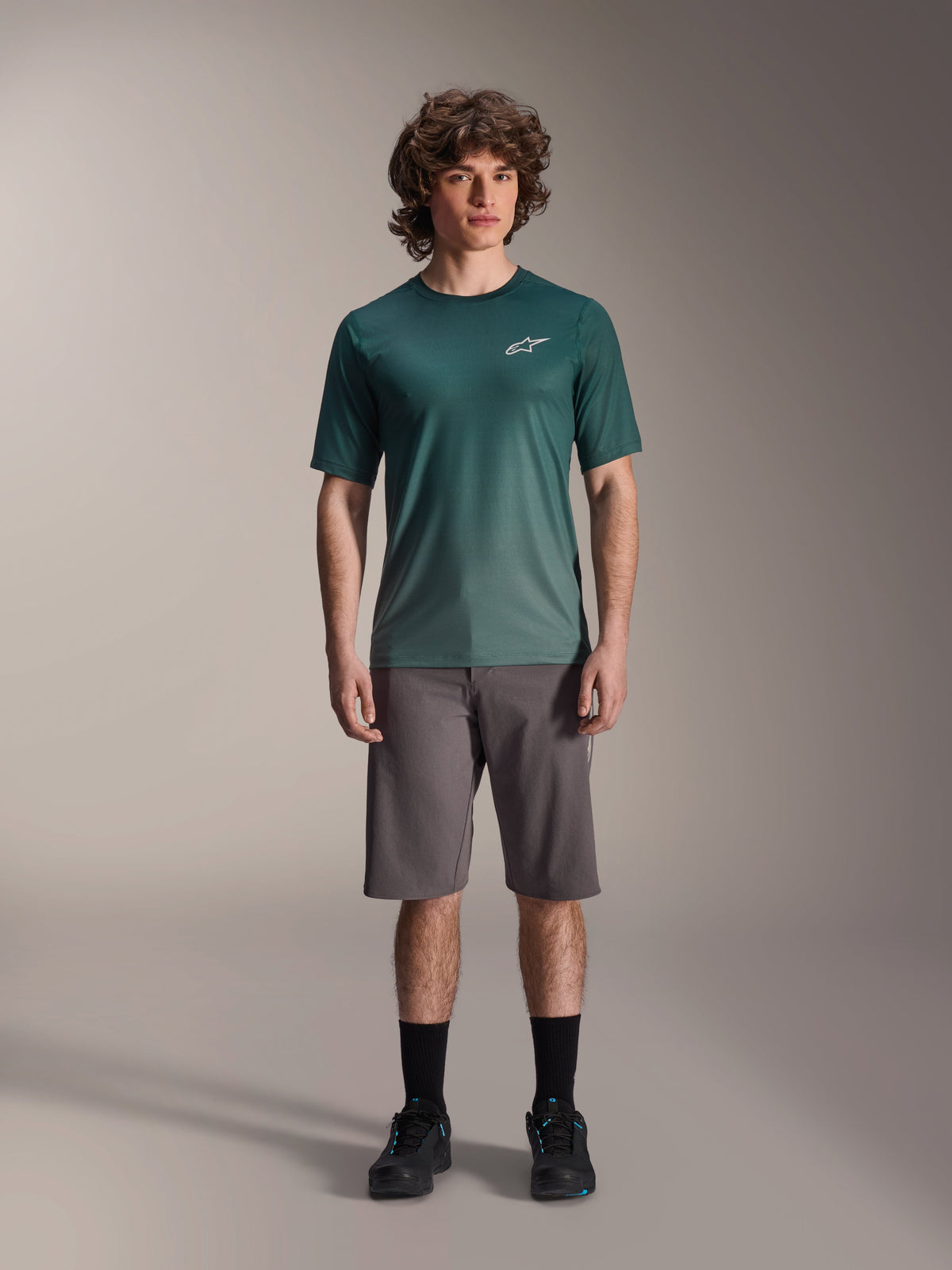 Male model wearing Alpinestars A-Dura Rise Jersey - Short Sleeve in Silver Pine, green cycling jersey with small white chest logo, paired with grey mountain bike shorts and black footwear, casual MTB trail gear