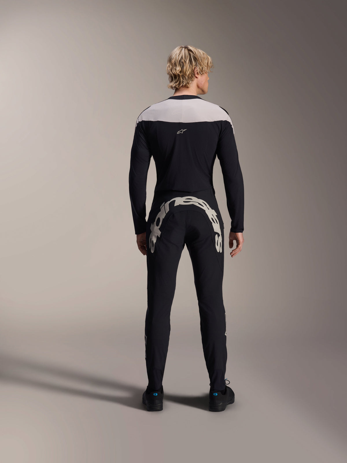 Alpinestars A-Supra Melt Pants, Black, back view showing the large white Alpinestars wordmark across the back yoke, paired with a matching long-sleeve jersey and black riding shoes, high-performance mountain bike gear for off-road riding.