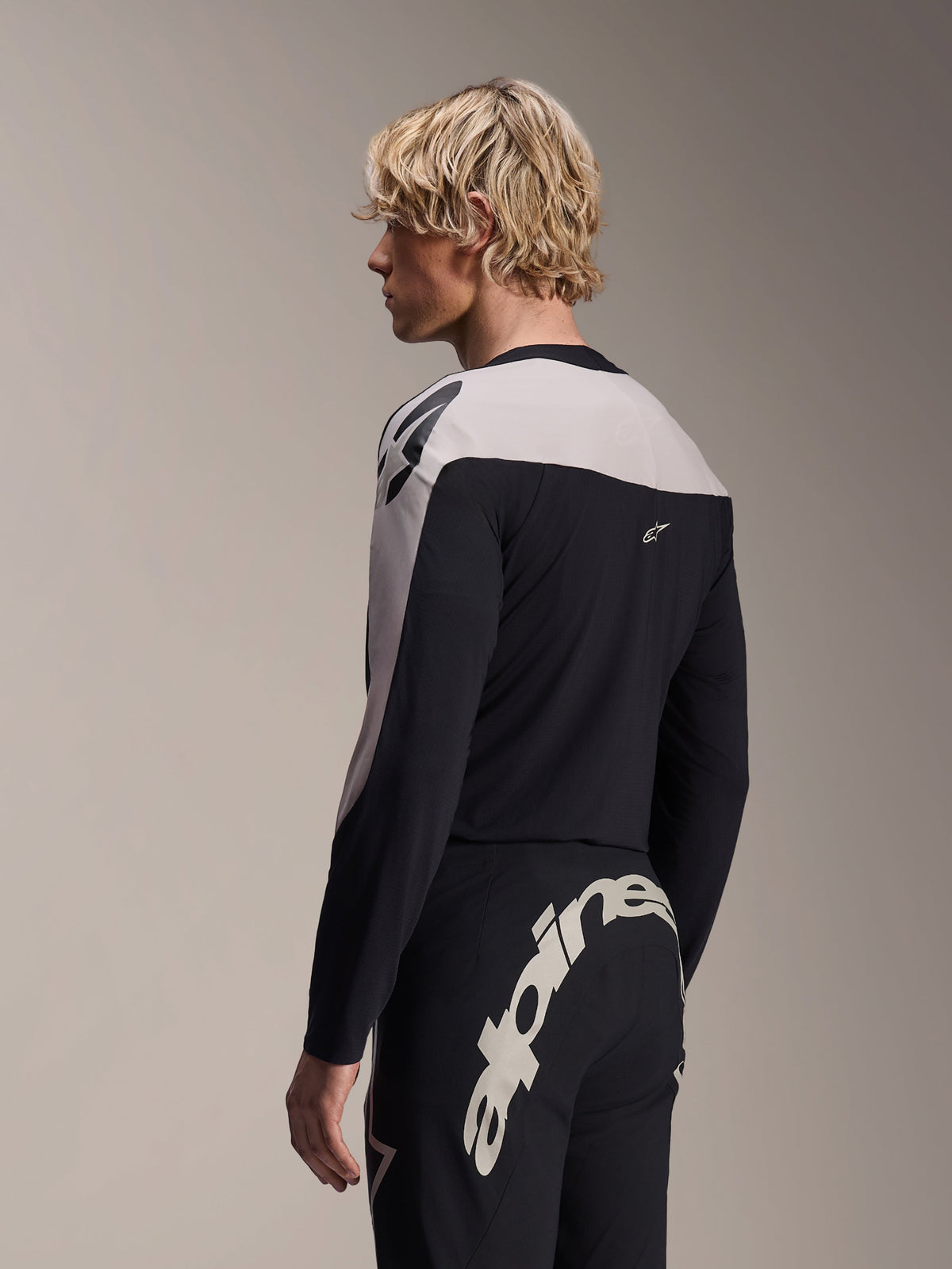 Male model wearing Alpinestars A-Supra Melt Long Sleeve Jersey and A-Supra Melt Pants, black and light grey MTB racing kit, featuring large bold white Alpinestars wordmark across the back yoke of the pants