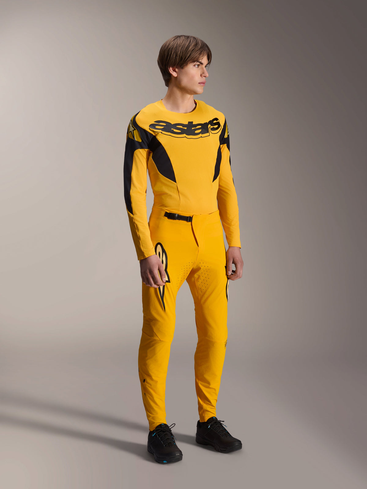 Modèle portant le maillot Alpinestars Limited Edition A-Supra Melt Gold, maillot de VTT à manches longues jaune foncé et noir, avec pantalon jaune assorti et chaussures noires, équipement technique de cyclisme tout-terrain aux graphismes inspirés de la course