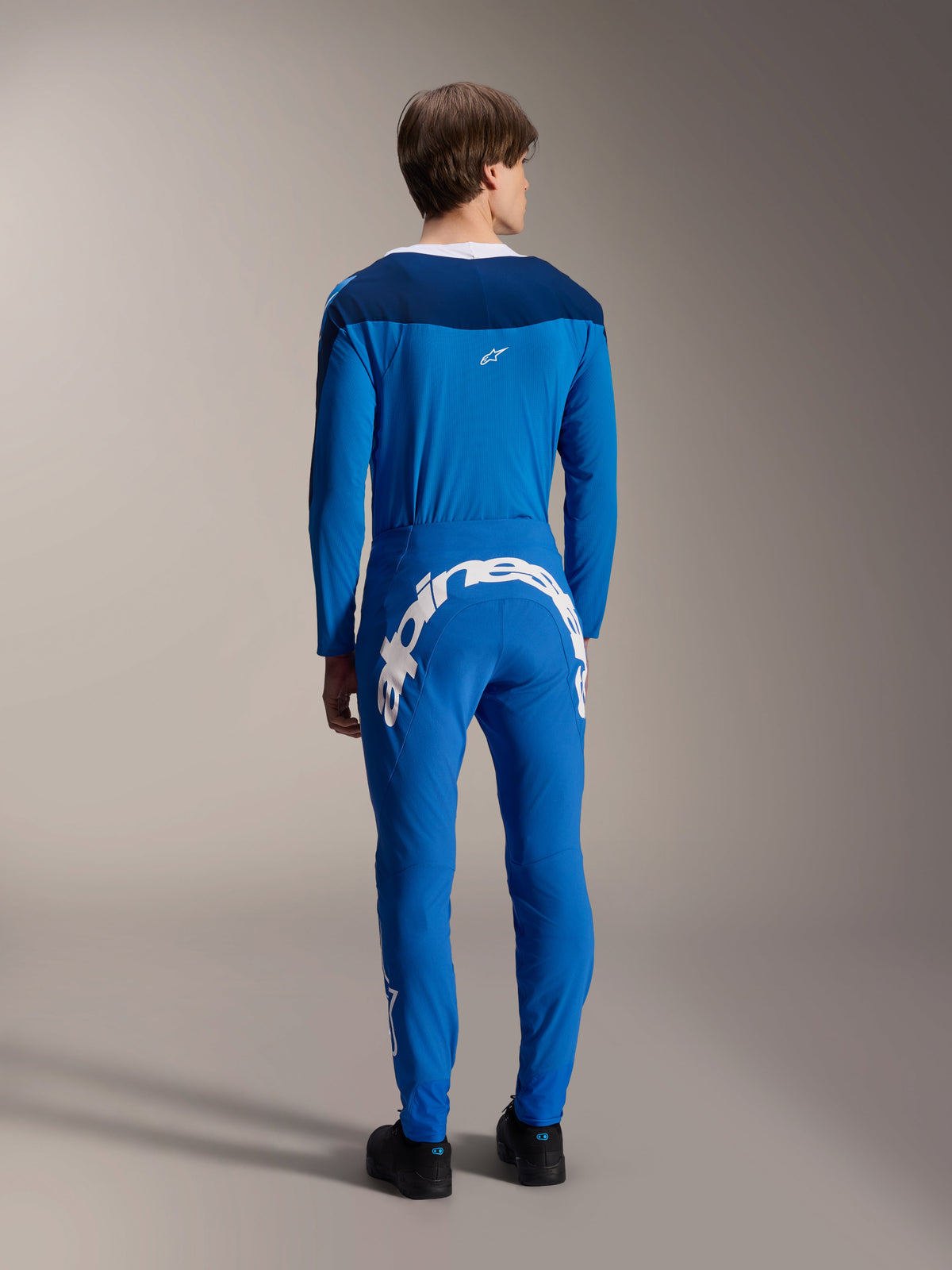 Modèle masculin vu de dos portant le maillot à manches longues Alpinestars A-Supra Melt en bleu, associé à un pantalon de performance bleu assorti arborant un grand logo textuel Alpinestars blanc dans le dos, tenue de course VTT complète.