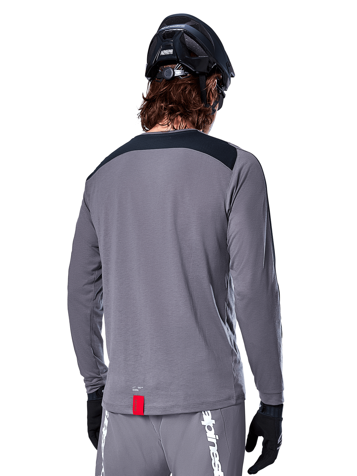 Maillot à manches longues Alpinestars A-Dura Dri Astar, maillot de VTT gris foncé vu de dos, avec empiècement noir aux épaules et petite étiquette rouge de la marque, porté avec un casque de VTT et des gants noirs.