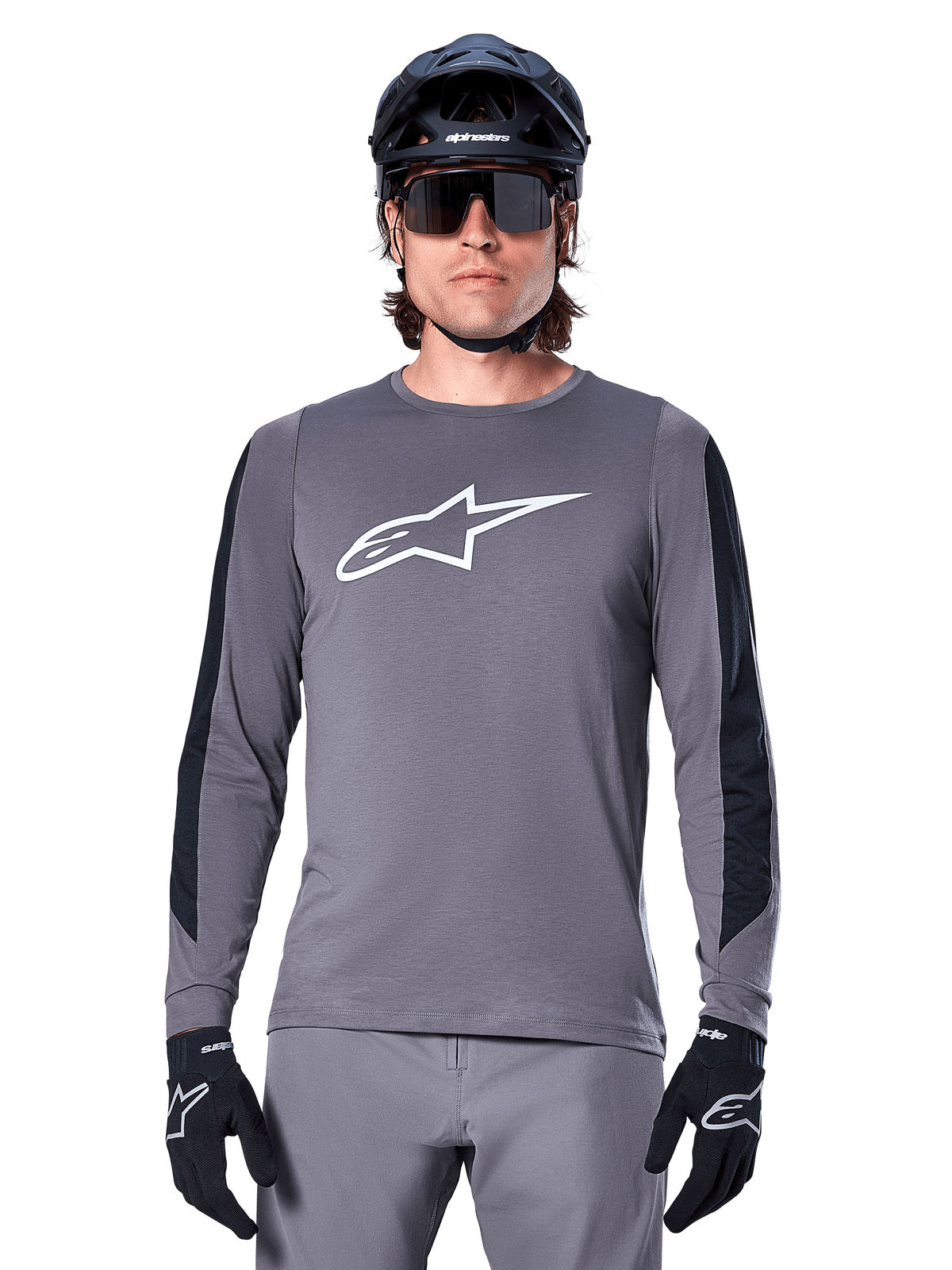 Maillot à manches longues Alpinestars A-Dura Dri Astar, gris foncé, avec un large logo Astar blanc sur la poitrine et des panneaux en maille noire sur les bras, porté par un cycliste avec un casque de VTT noir et des lunettes de soleil de performance, équipement de cyclisme VTT