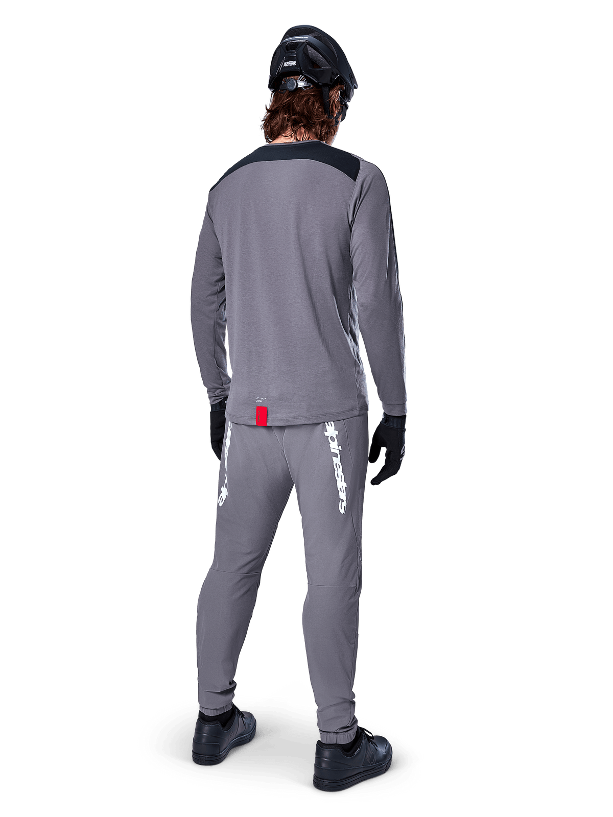 Maillot Alpinestars A-Dura Dri Astar à manches longues, gris foncé, vue de dos d'un cycliste portant un pantalon assorti et un casque noir, avec empiècements noirs aux épaules et une petite étiquette rouge à l'ourlet, équipement technique de cyclisme tout-terrain