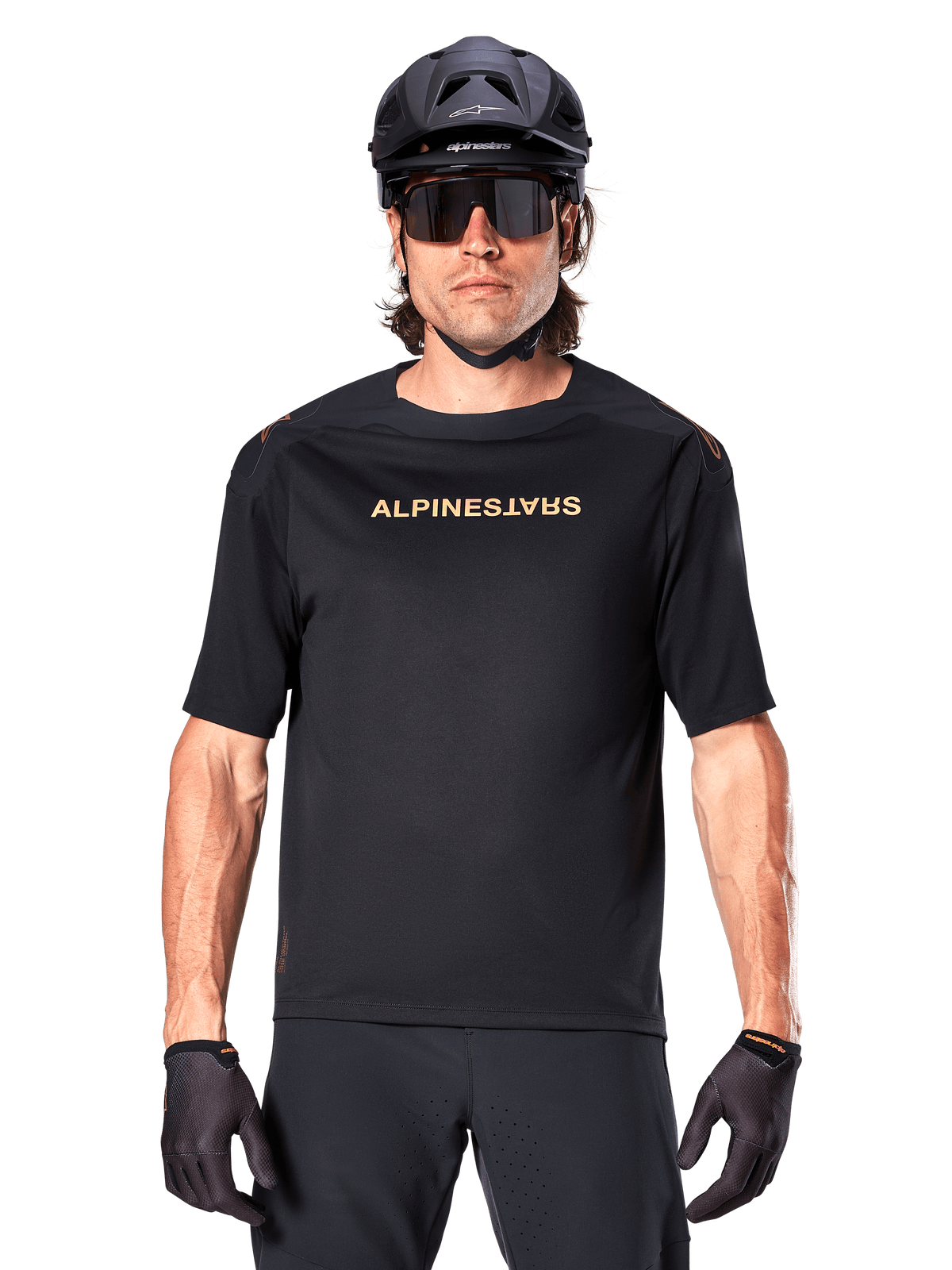 Vététiste portant un maillot à manches courtes Alpinestars A-Aria Polartec Switch noir, avec logo doré sur la poitrine, accompagné d'un casque de VTT noir, de lunettes de soleil de sport sombres et de gants assortis, équipement de cyclisme tout-terrain