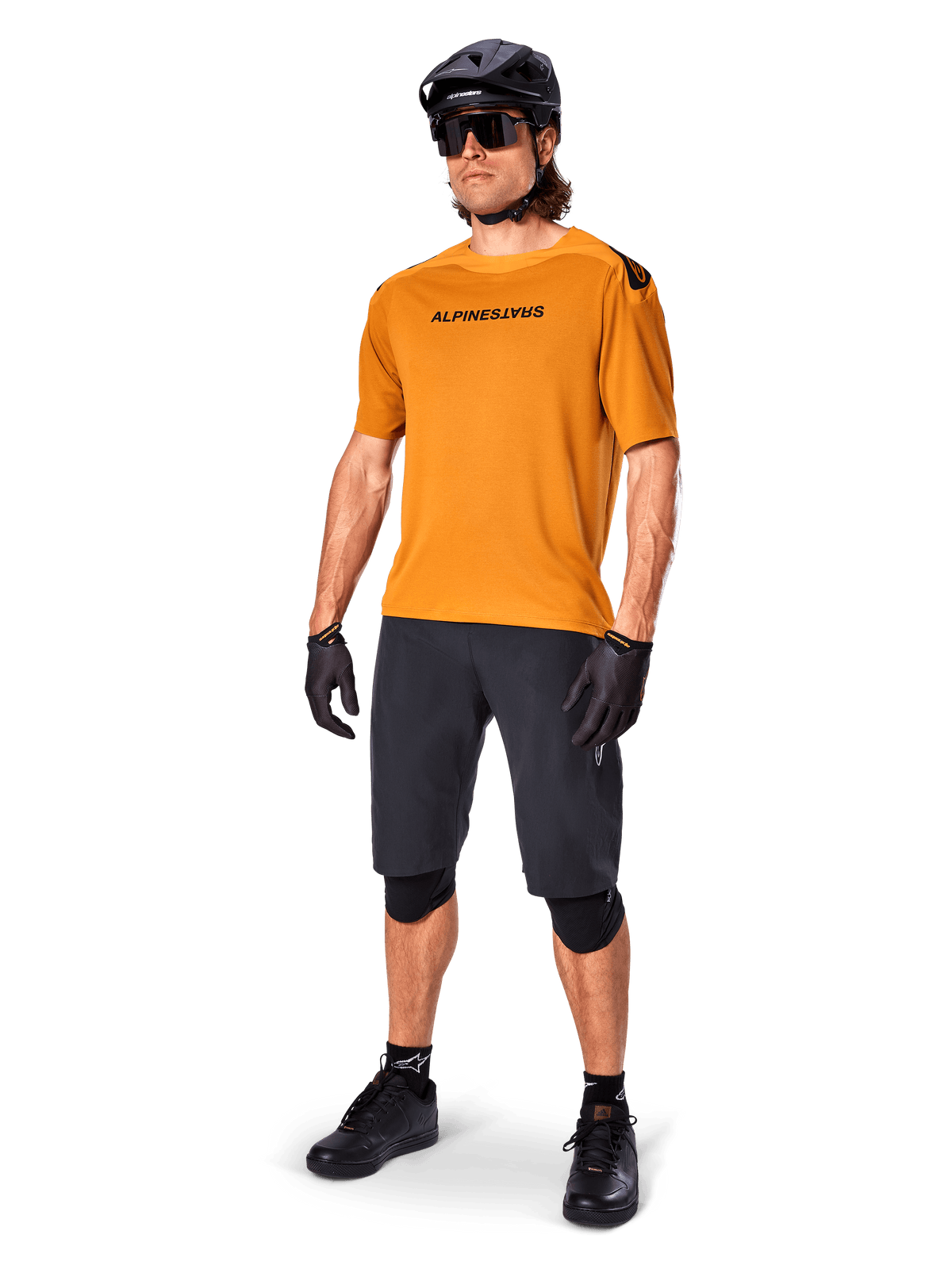 Cycliste portant le maillot Alpinestars A-Aria Polartec Switch en Dark Gold, maillot de VTT à manches courtes, associé à un short de cyclisme noir, des genouillères, un casque noir et des gants, tenue de VTT professionnelle pour la pratique du trail