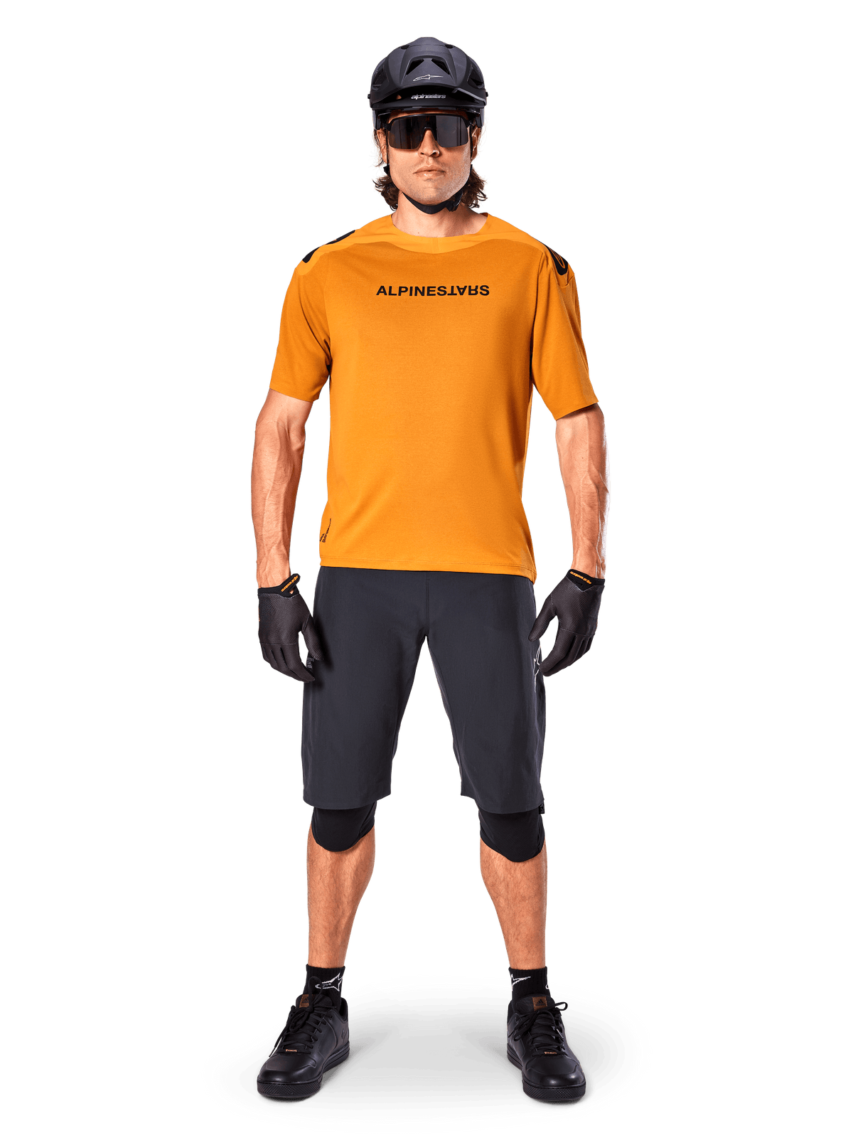 Cycliste portant le maillot à manches courtes Alpinestars A-Aria Polartec Switch en Or Foncé, assorti d'un short de VTT noir, d'un casque, de lunettes de soleil et de gants, équipement professionnel pour le VTT de trail
